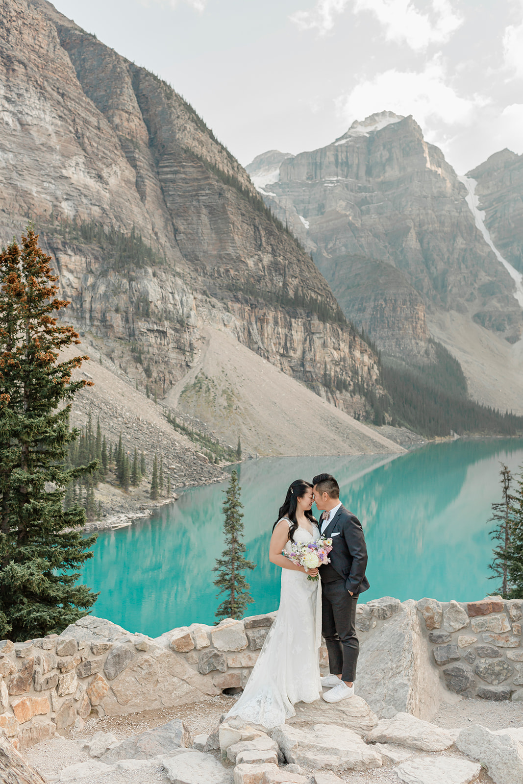 A couple in formal wedding attire embraces on an overlook at Moraine Lake in Banff National Park.