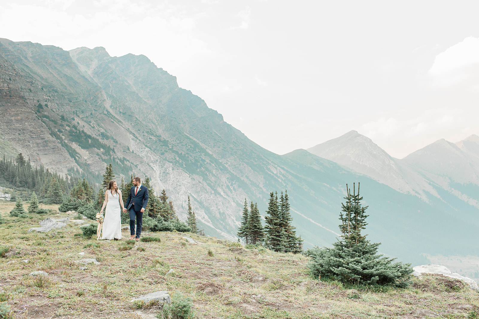 A Banff National Park elopement couple walks along a meadow that overlooks a valley surrounded by mountains. 