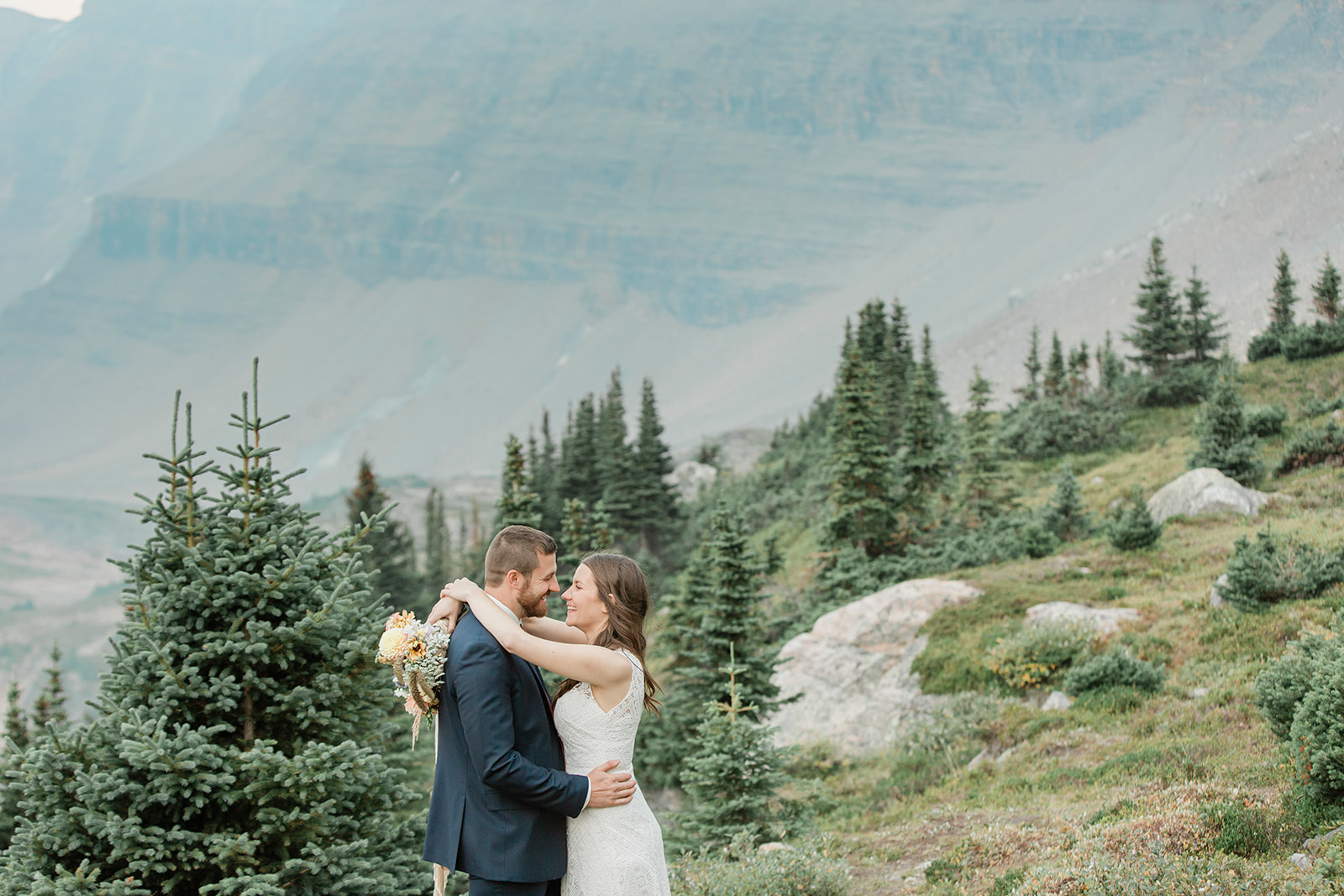 A Banff National Park elopement couple embraces on a hillside with evergreen trees in the background. 