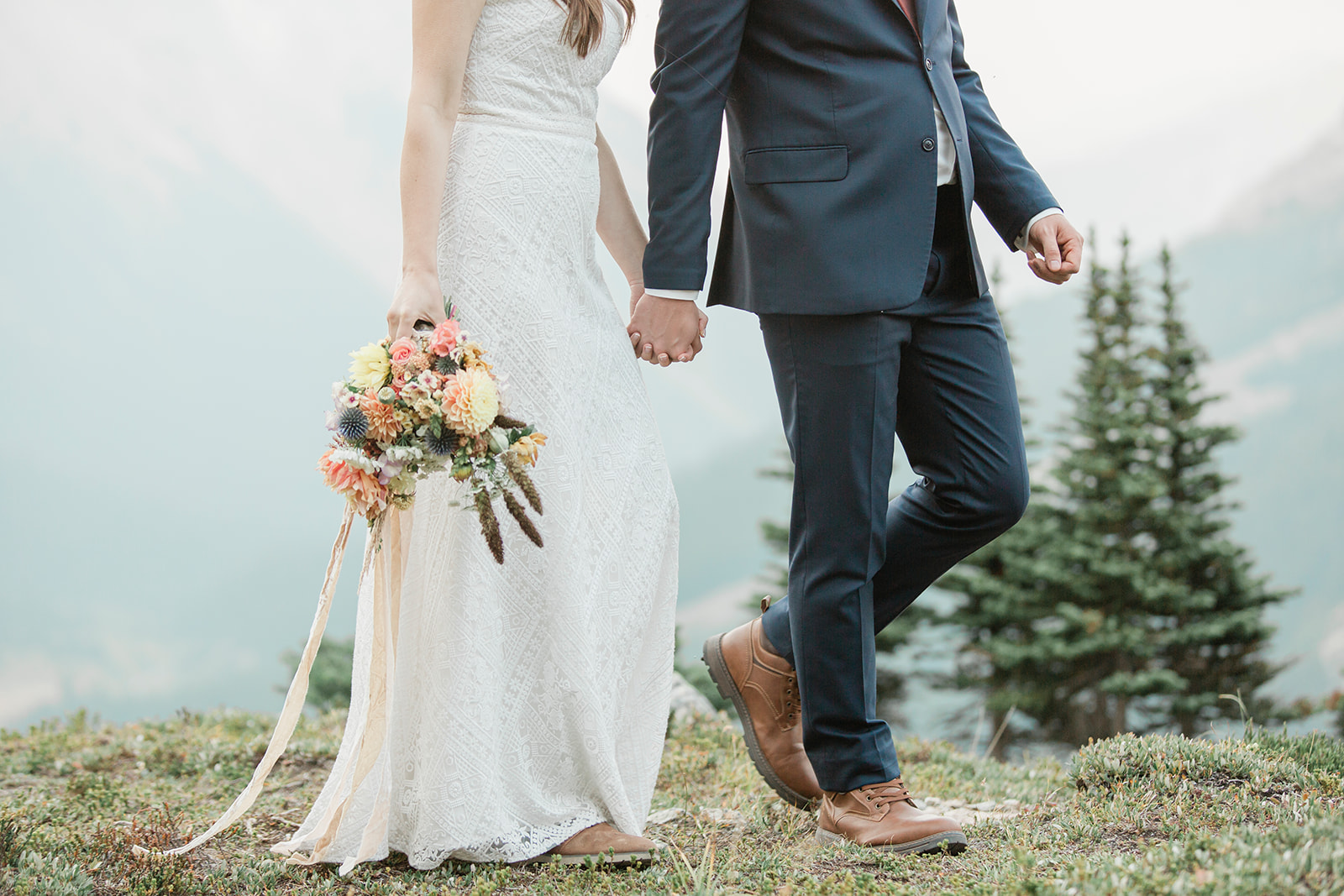 A woman in a long white wedding gown holds a bright floral bouquet and her groom's hand while walking in Banff National Park. 