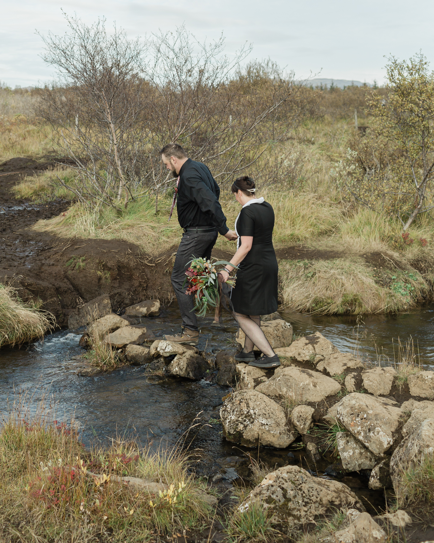 Before their Iceland elopement ceremony, Virginia and Derek are walking over a creek in front of a mountain in the Golden Circle
