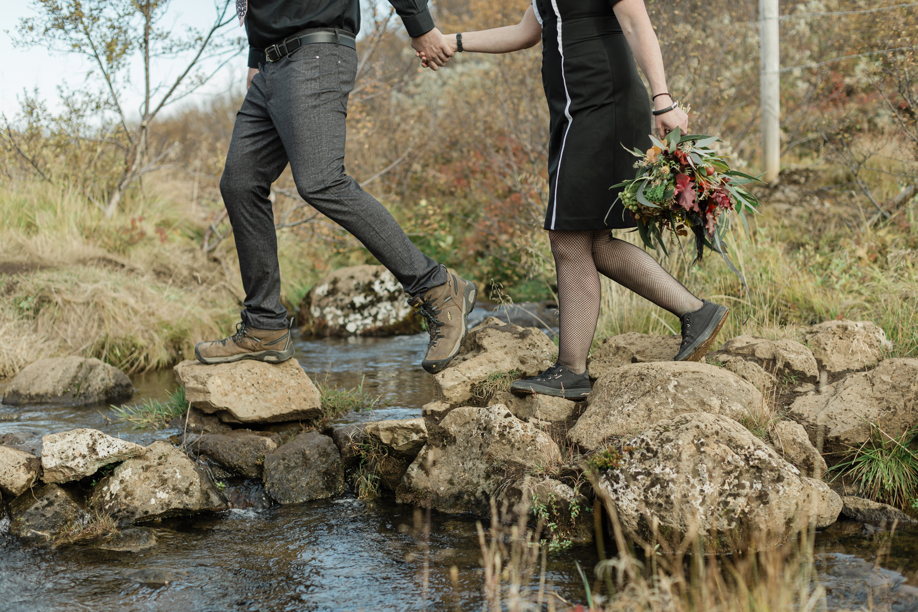A couple that are walking hand in hand, passing over rocks and water, towards their waterfall ceremony location