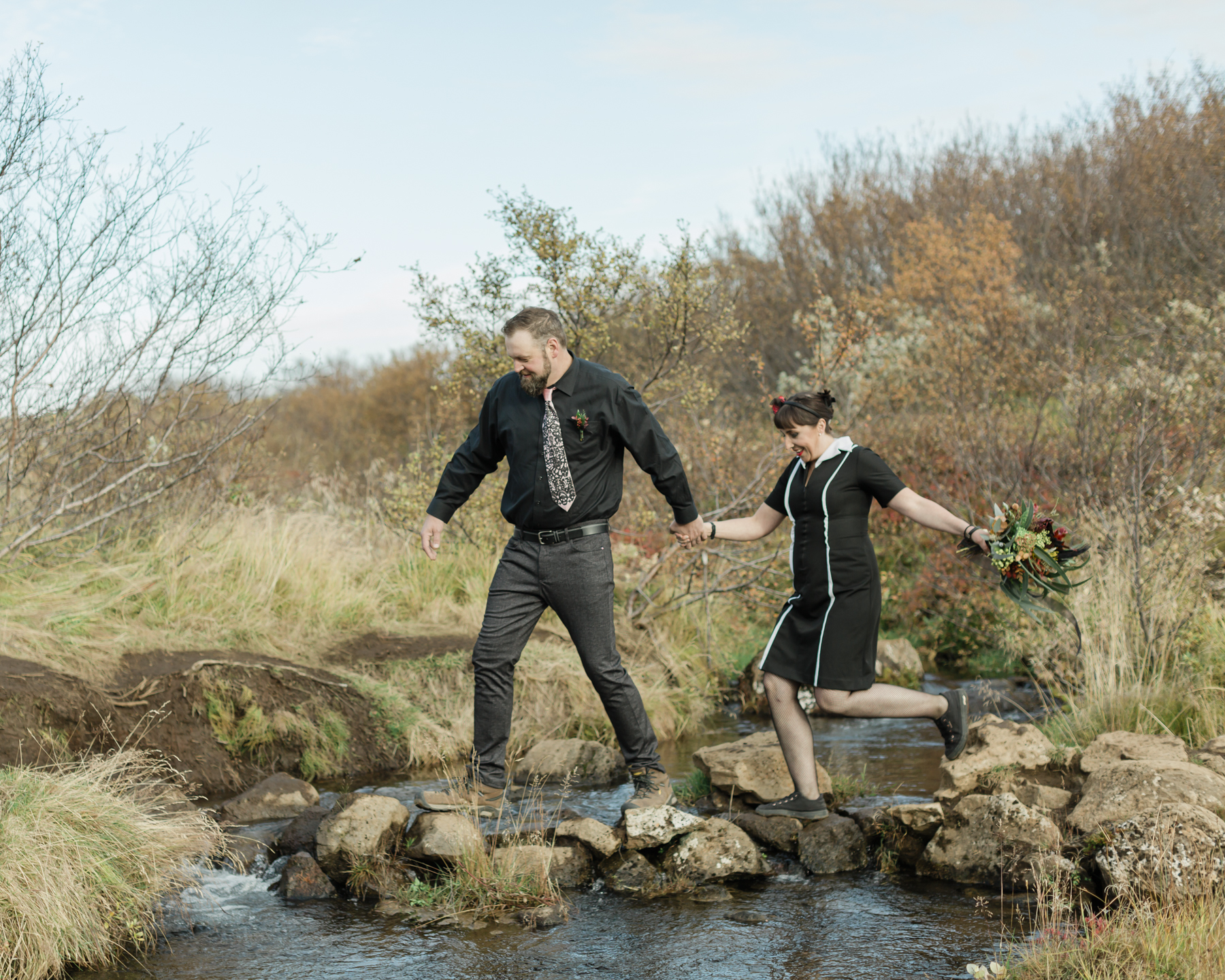 A couple that are walking hand in hand, passing over rocks and water, towards their waterfall ceremony location
