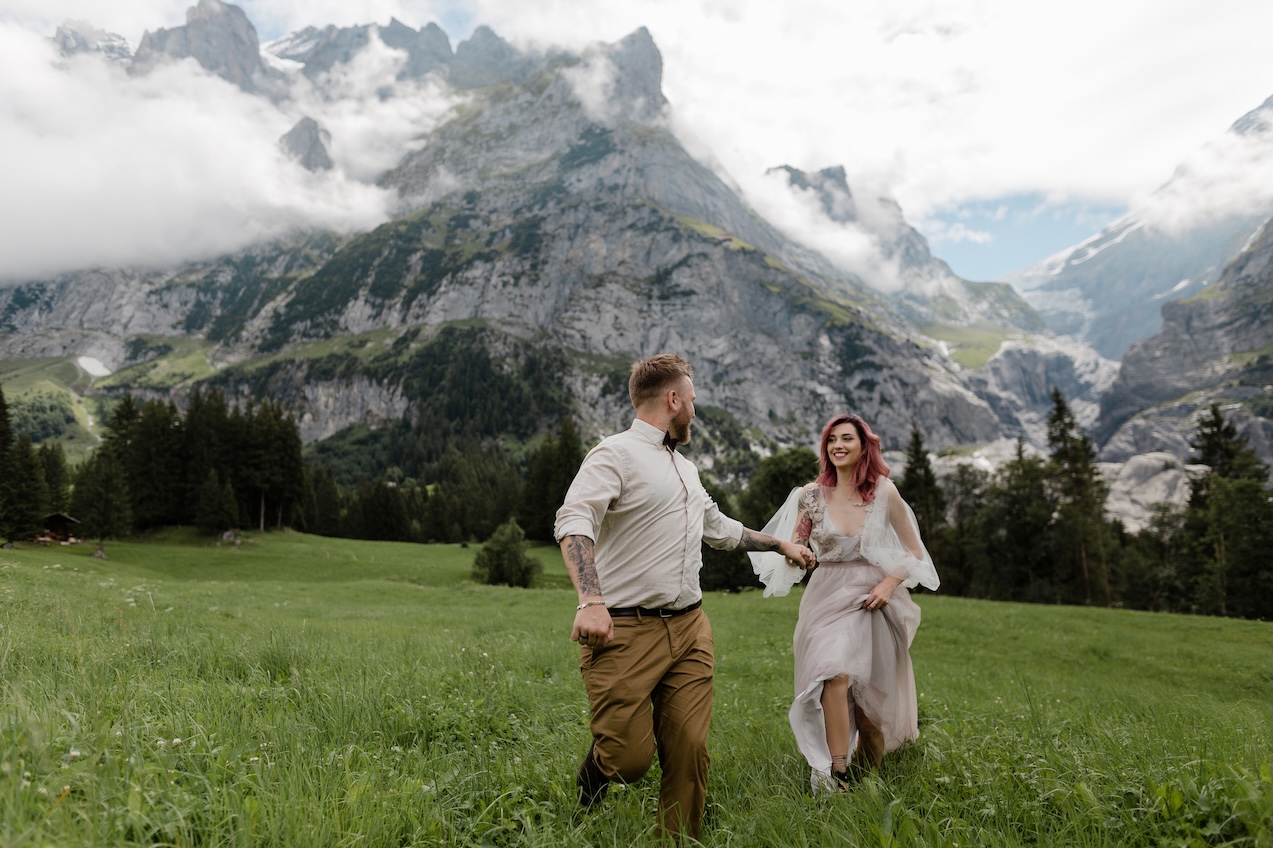 happy bride in wedding dress and groom holding hands and walking on alpine meadow under Blüemlisalp Massif
