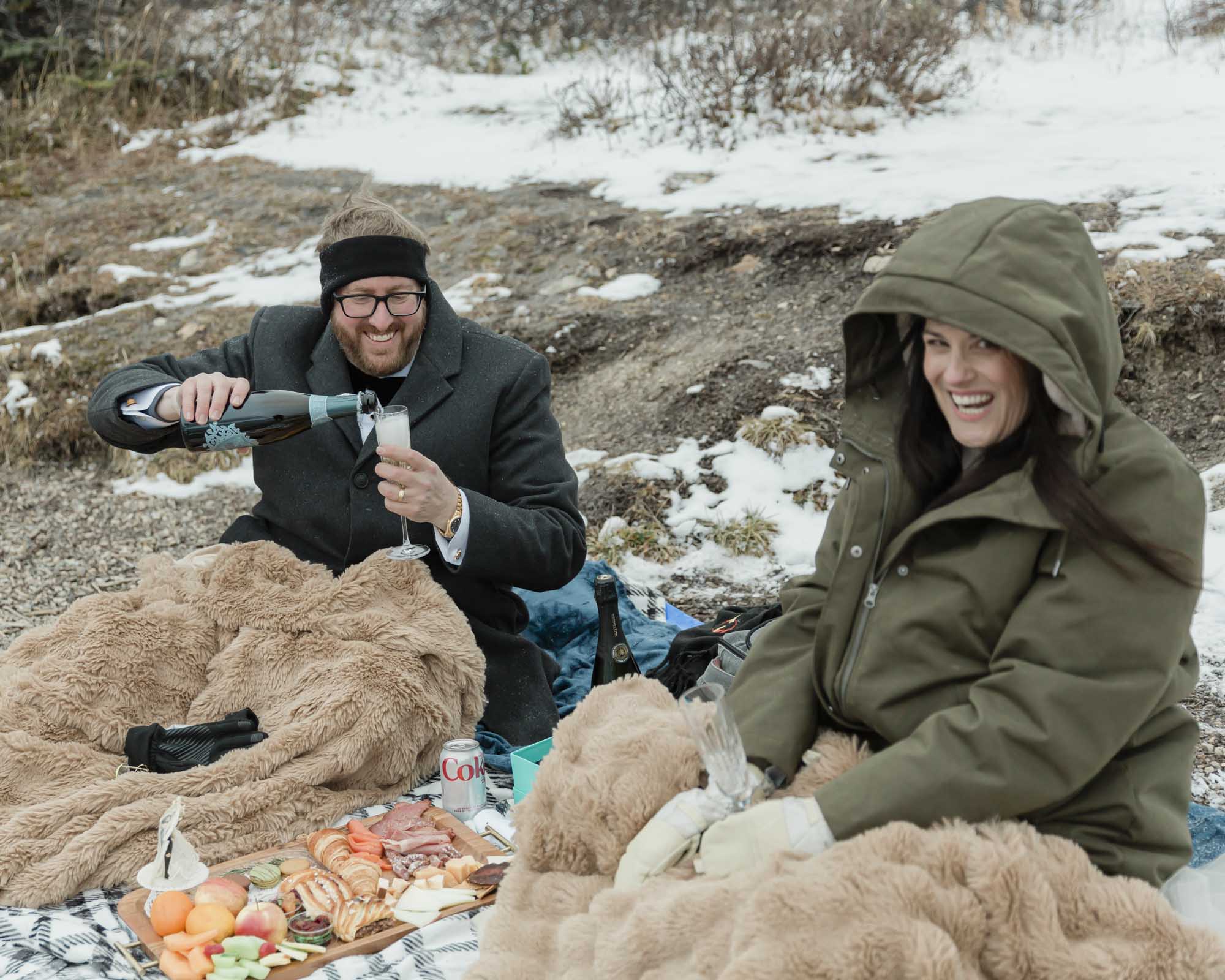 Wedding couple enjoying a winter picnic at Bow Lake in Banff National Park during their Elopement, featuring diet coke and a charcuterie board