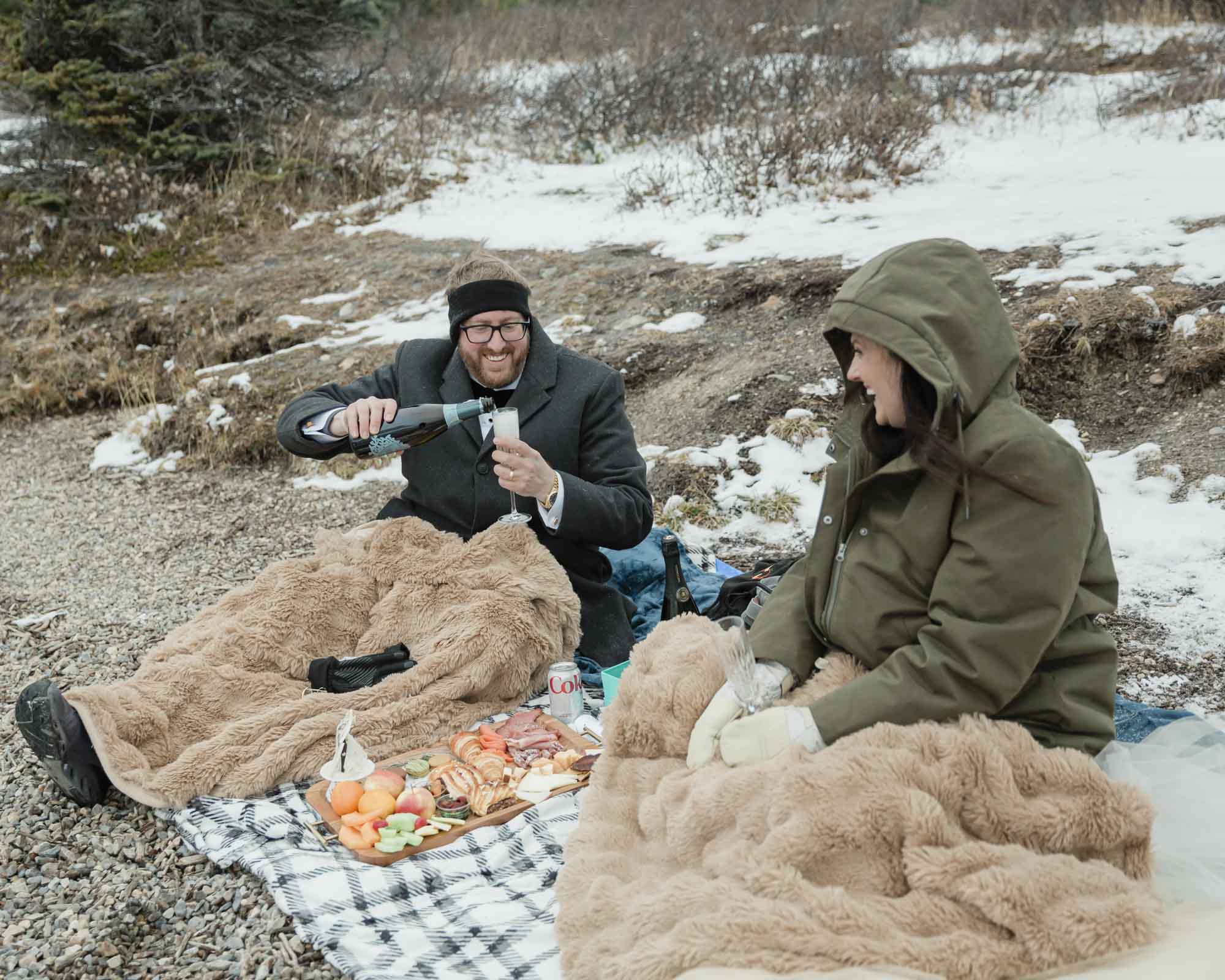 Wedding couple enjoying a winter picnic at Bow Lake in Banff National Park during their Elopement, featuring diet coke and a charcuterie board