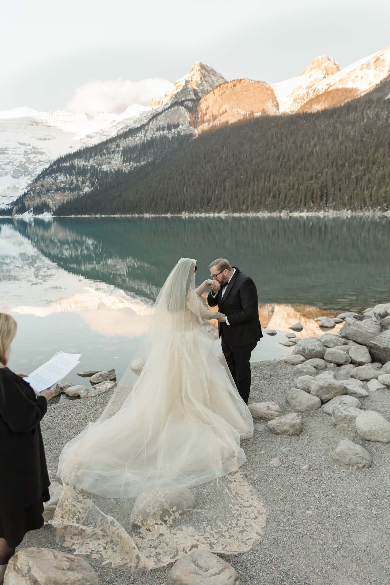a mountain elopement, the couple is holding hands in front of the reflective lake and the officiant is to their left guiding their ceremony.