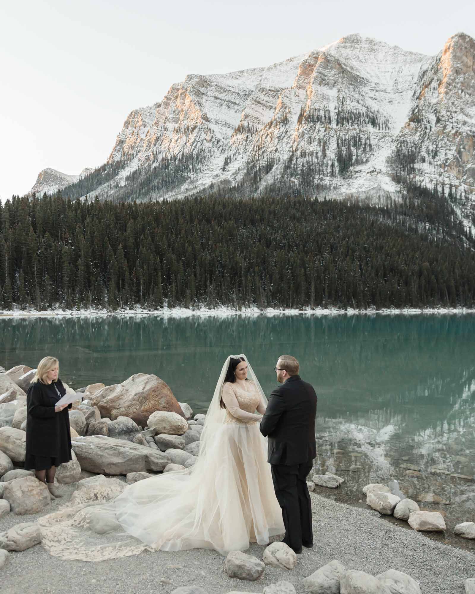 a mountain elopement, the couple is holding hands in front of the reflective lake and the officiant is to their left guiding their ceremony.