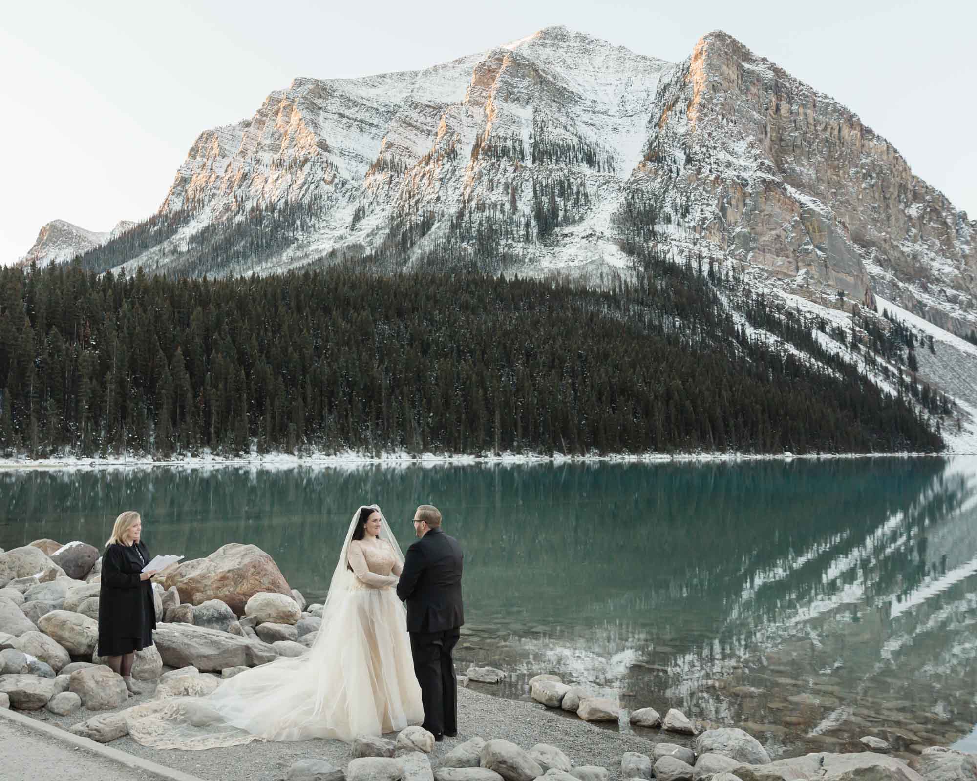 a mountain elopement, the couple is holding hands in front of the reflective lake and the officiant is to their left guiding their ceremony.