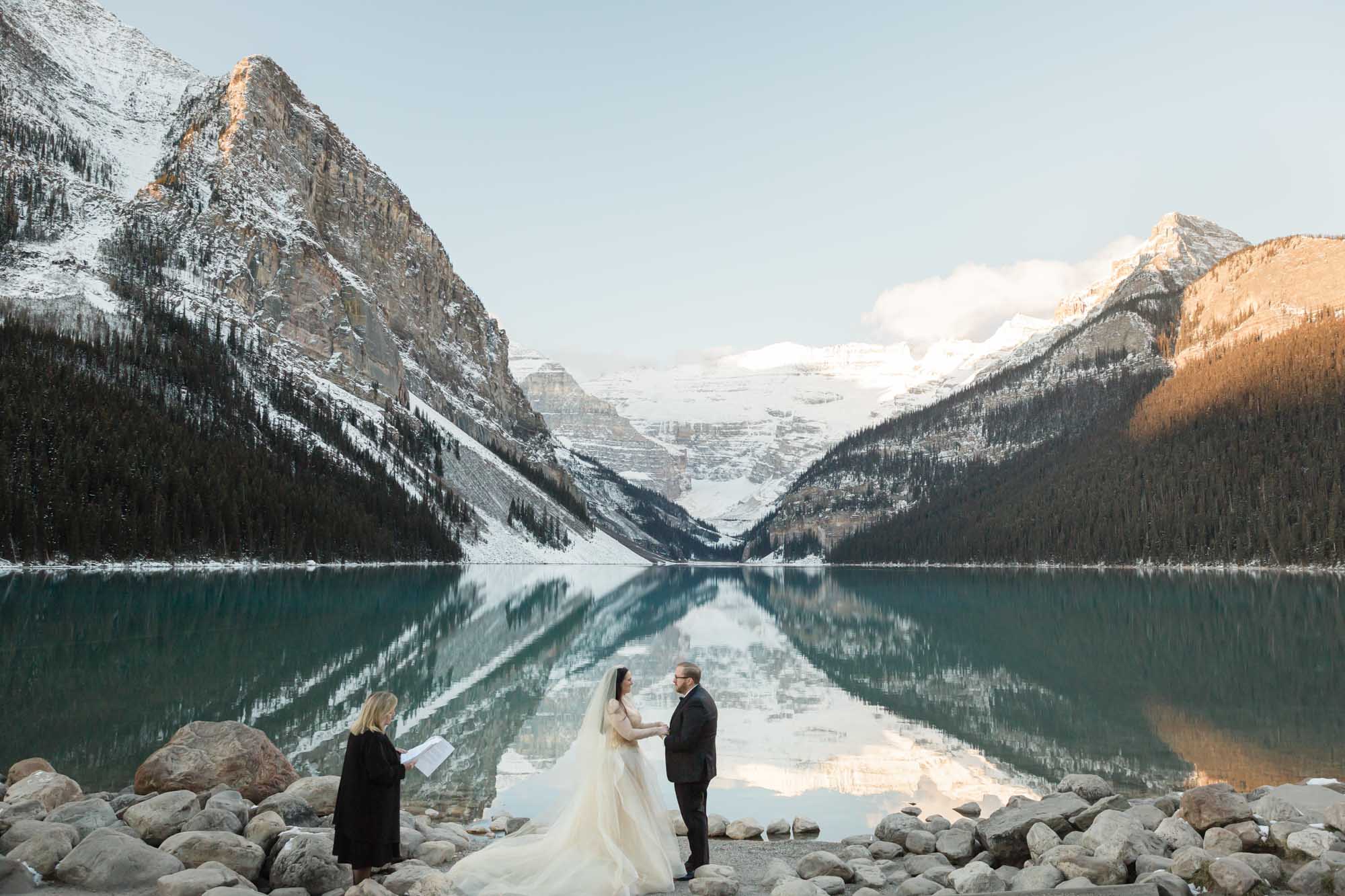 a mountain elopement, the couple is holding hands in front of the reflective lake and the officiant is to their left guiding their ceremony.