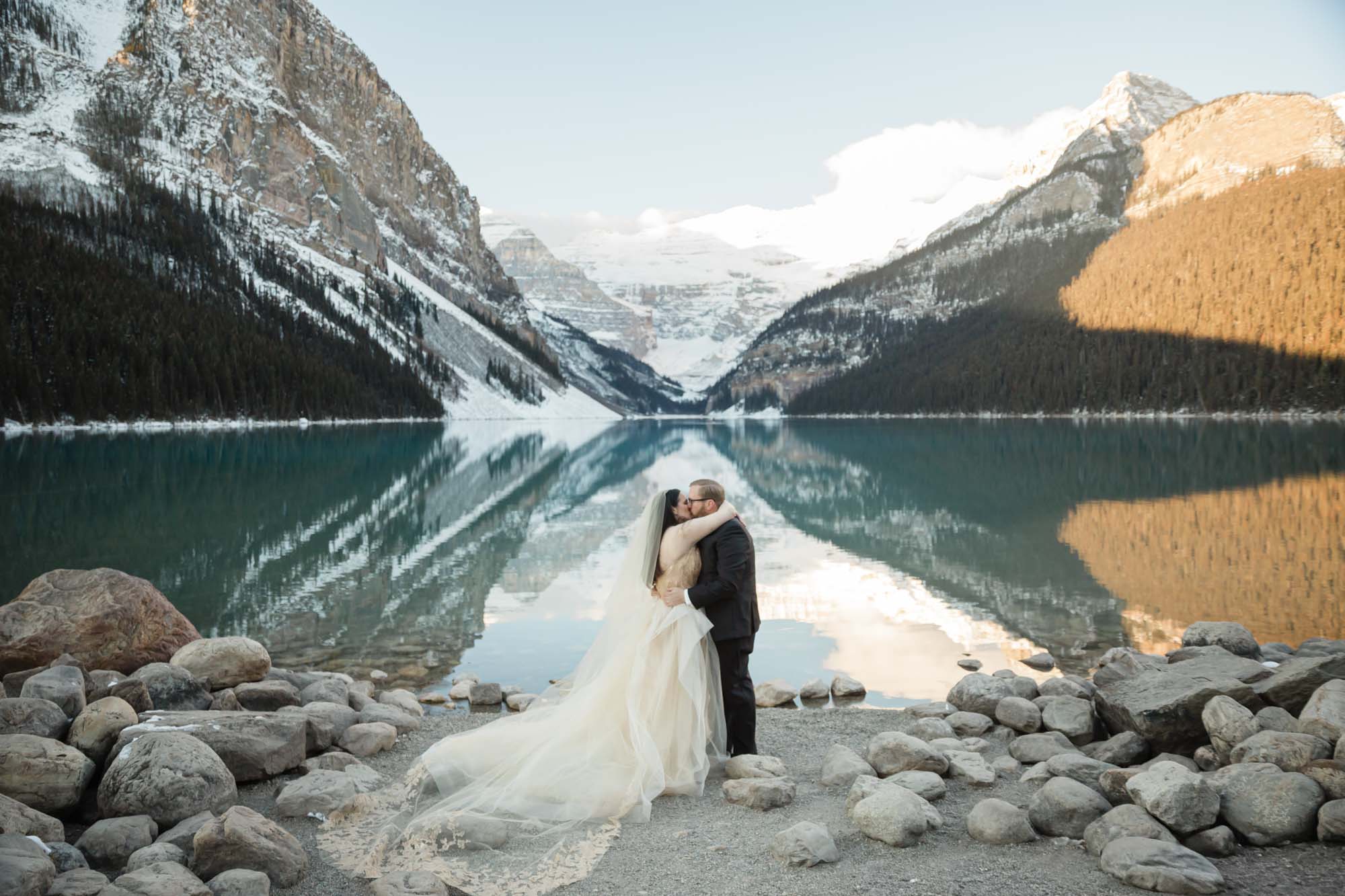 A Lake Louise elopement, the couple is holding hands in front of the reflective lake at sunrise in the winter