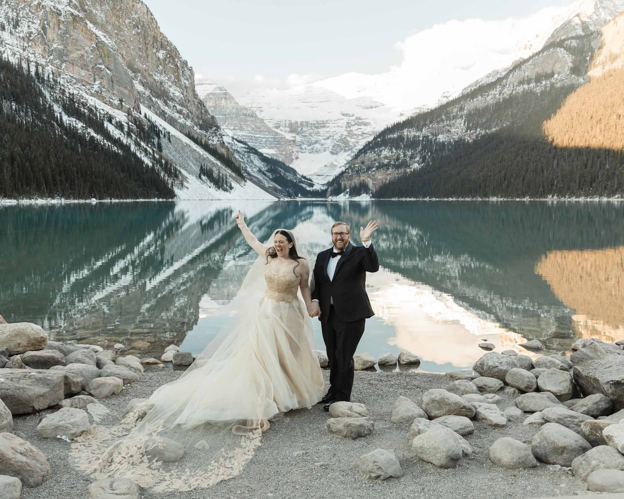 A Lake Louise elopement, the couple is holding hands in front of the reflective lake at sunrise in the winter