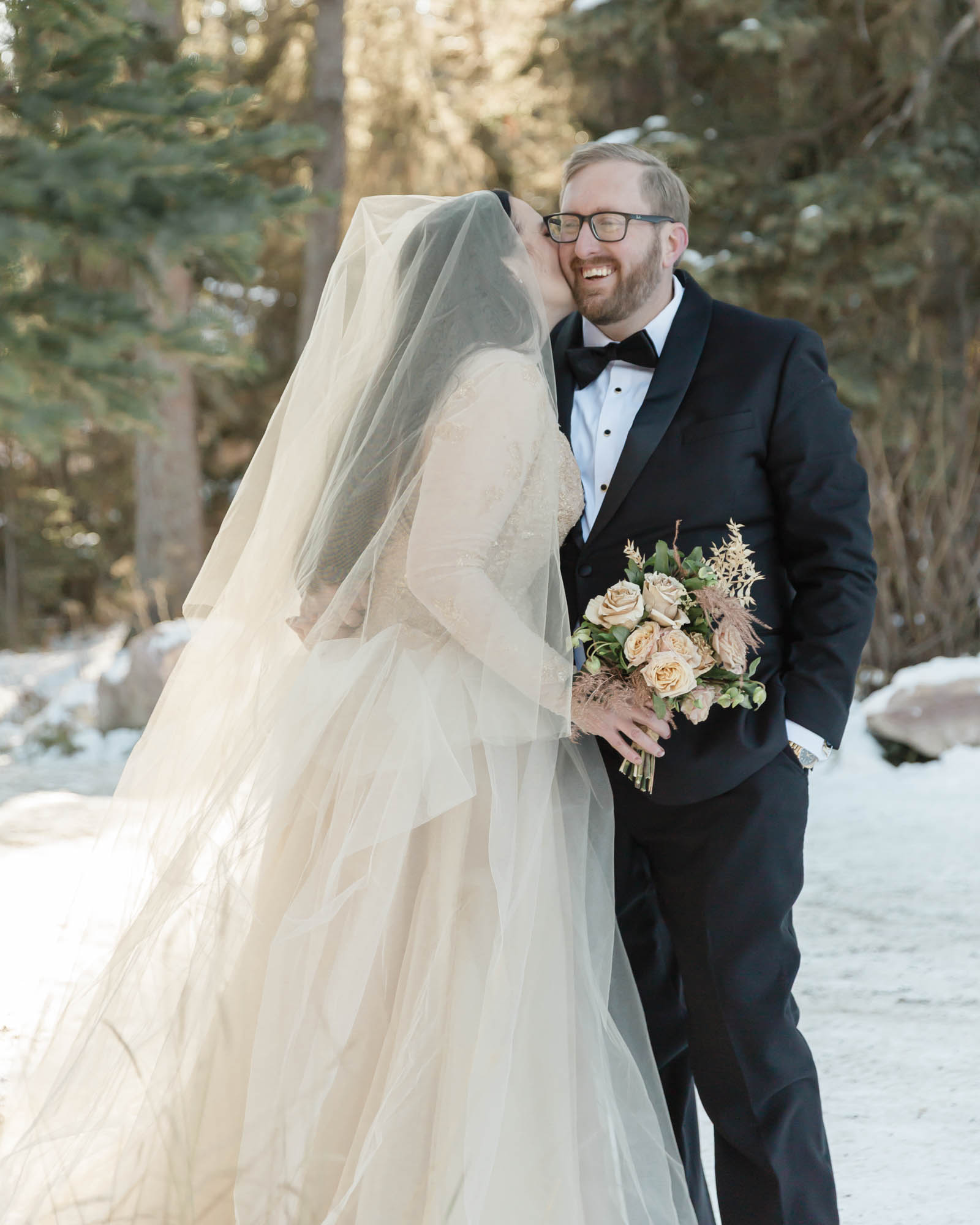 Wedding portraits in the forest in Banff National Park, the bride is kissing the grooms cheek