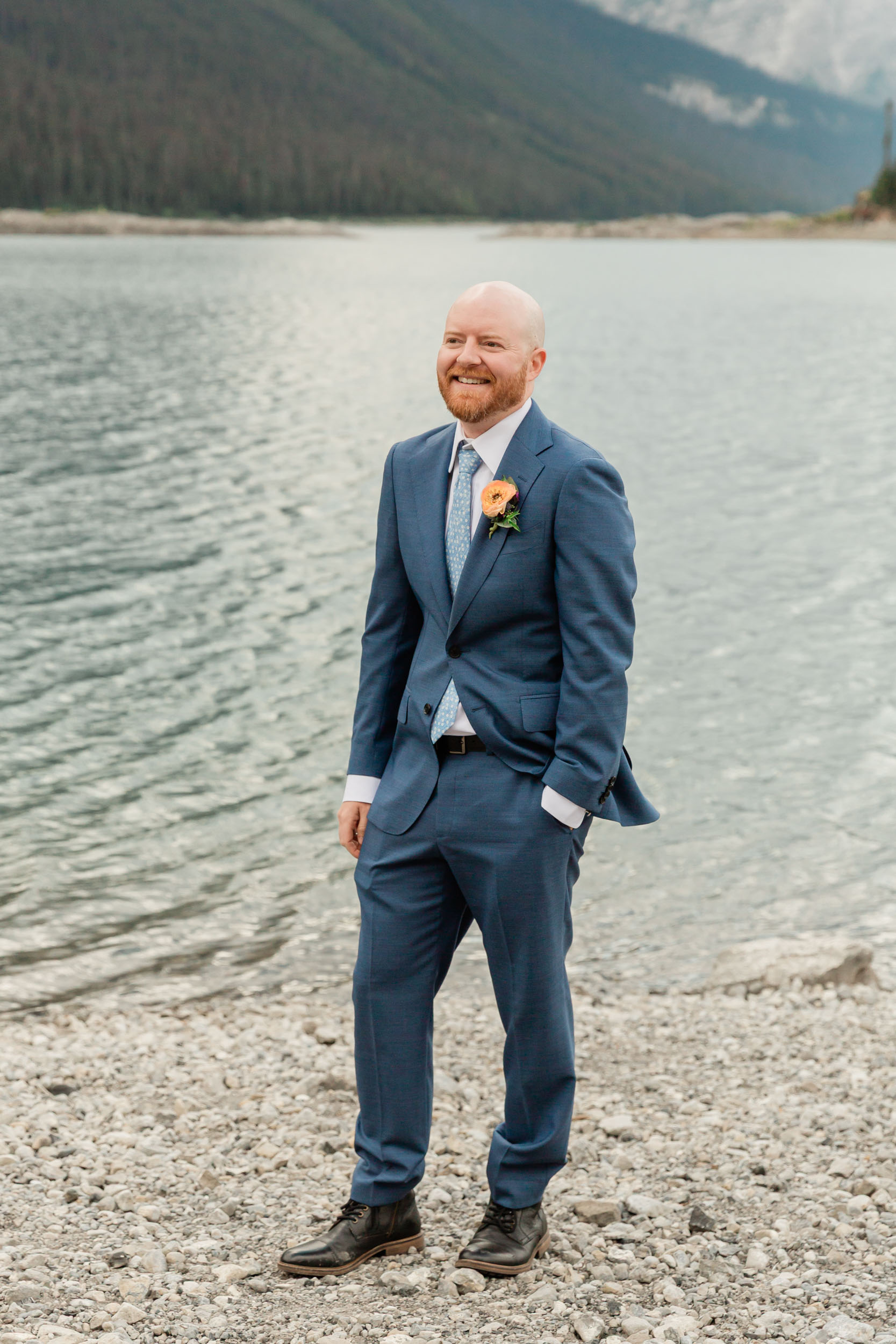 the groom is laughing during his portrait in front of the alpine lake and mountain views