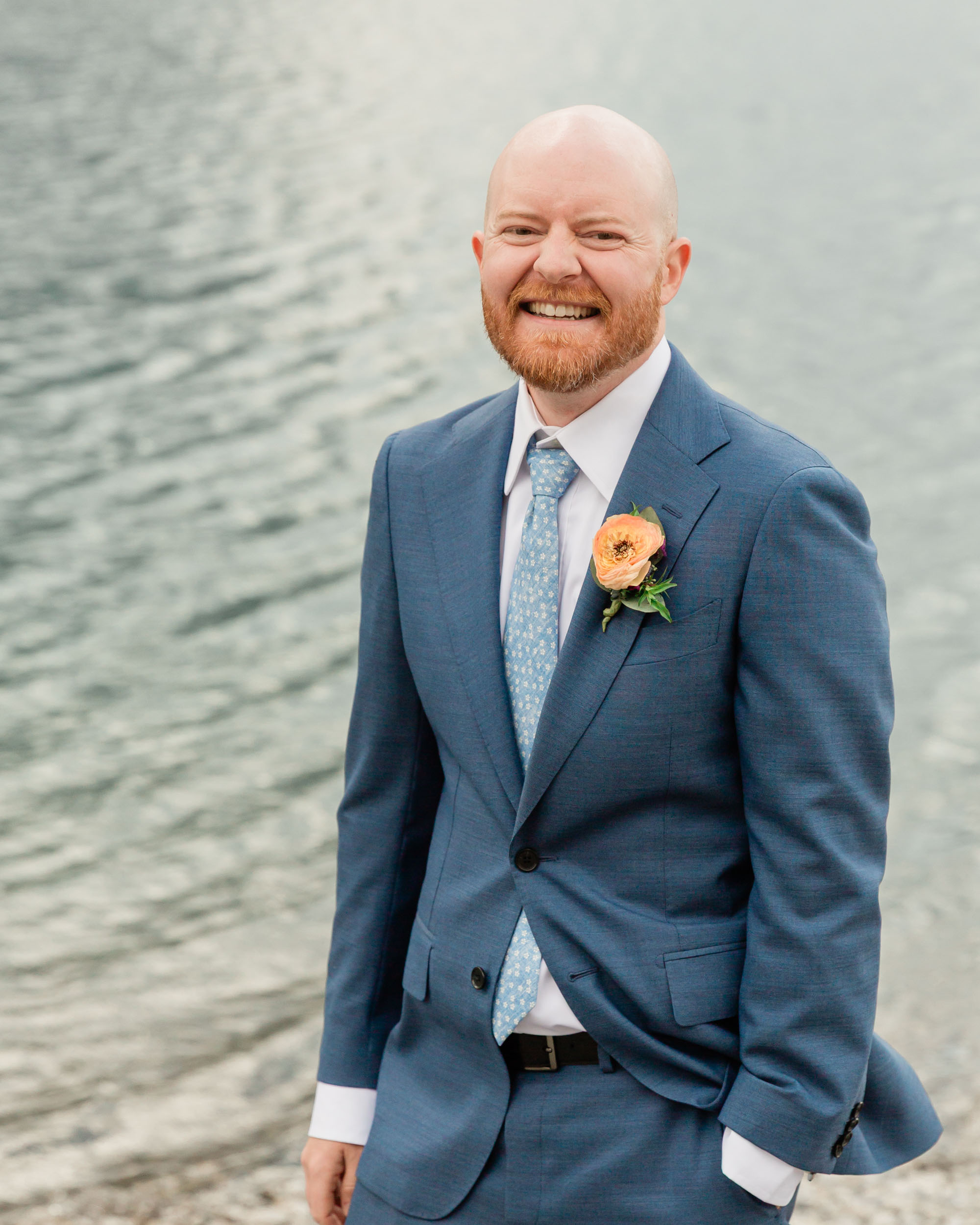 the groom is laughing during his portrait in front of the alpine lake and mountain views