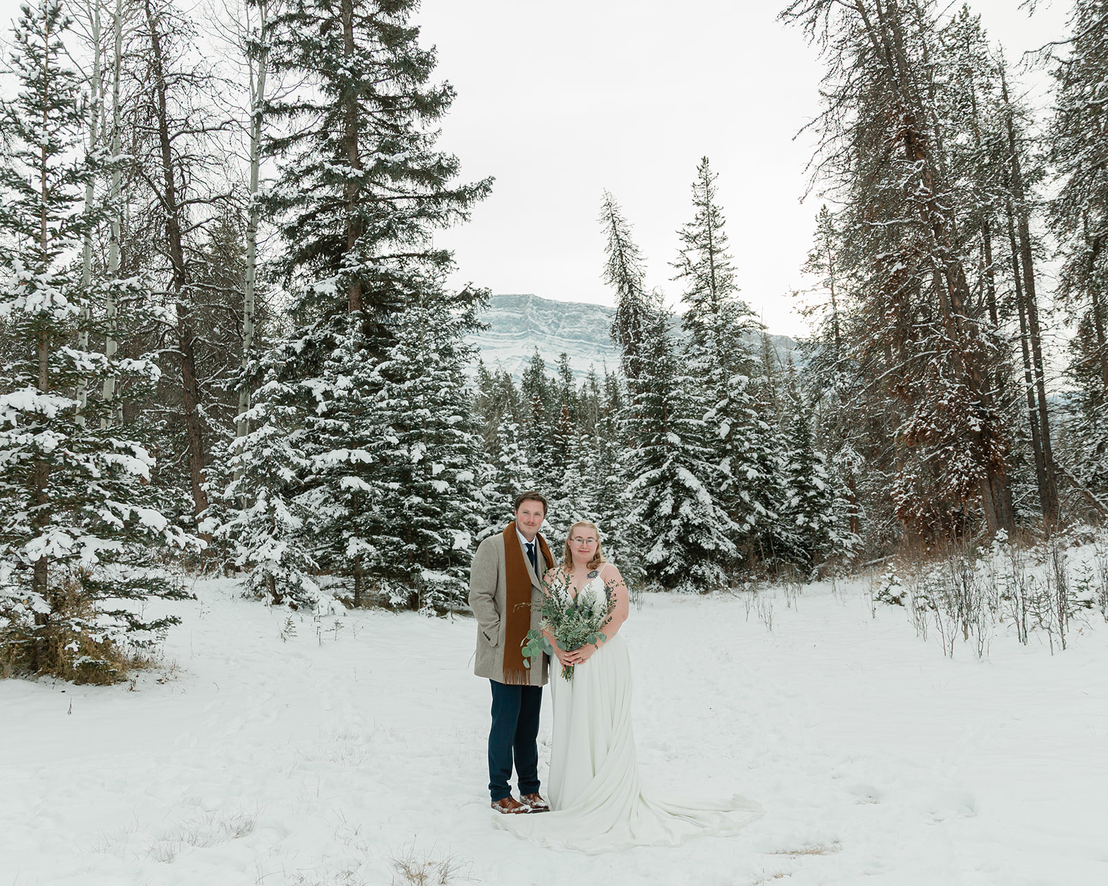 Couple posing together in a snowy forest with mountain views during their Banff winter elopement in Banff National Park.