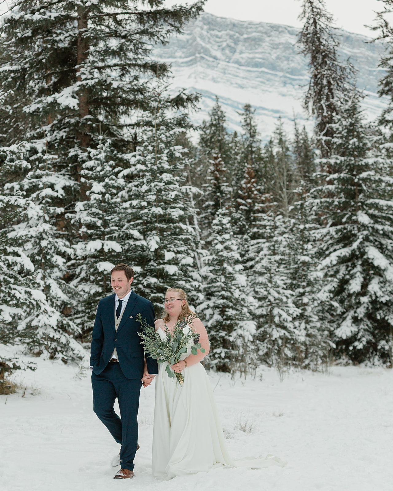 Newlywed couple walking hand in hand through a snowy mountain forest during their Banff winter elopement in Banff National Park.