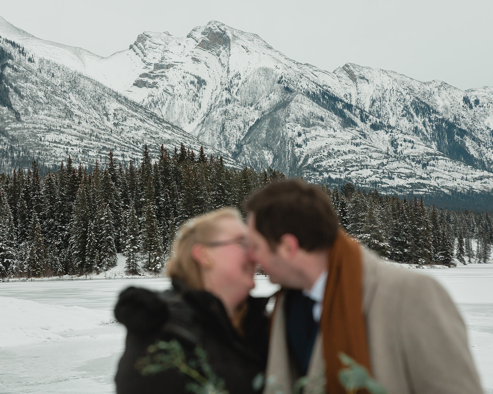 Gabriella and Bronson kiss in front of snow-covered Rocky Mountains during their romantic Banff winter elopement in Alberta.