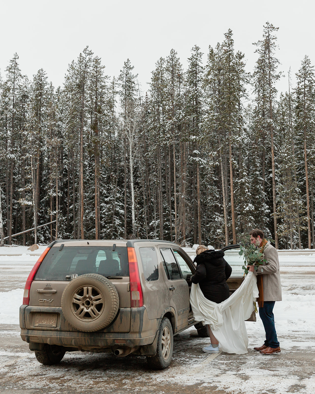 Gabriella and Bronson get into their car while carrying her bouquet and wedding dress during their snowy Banff winter elopement adventure.