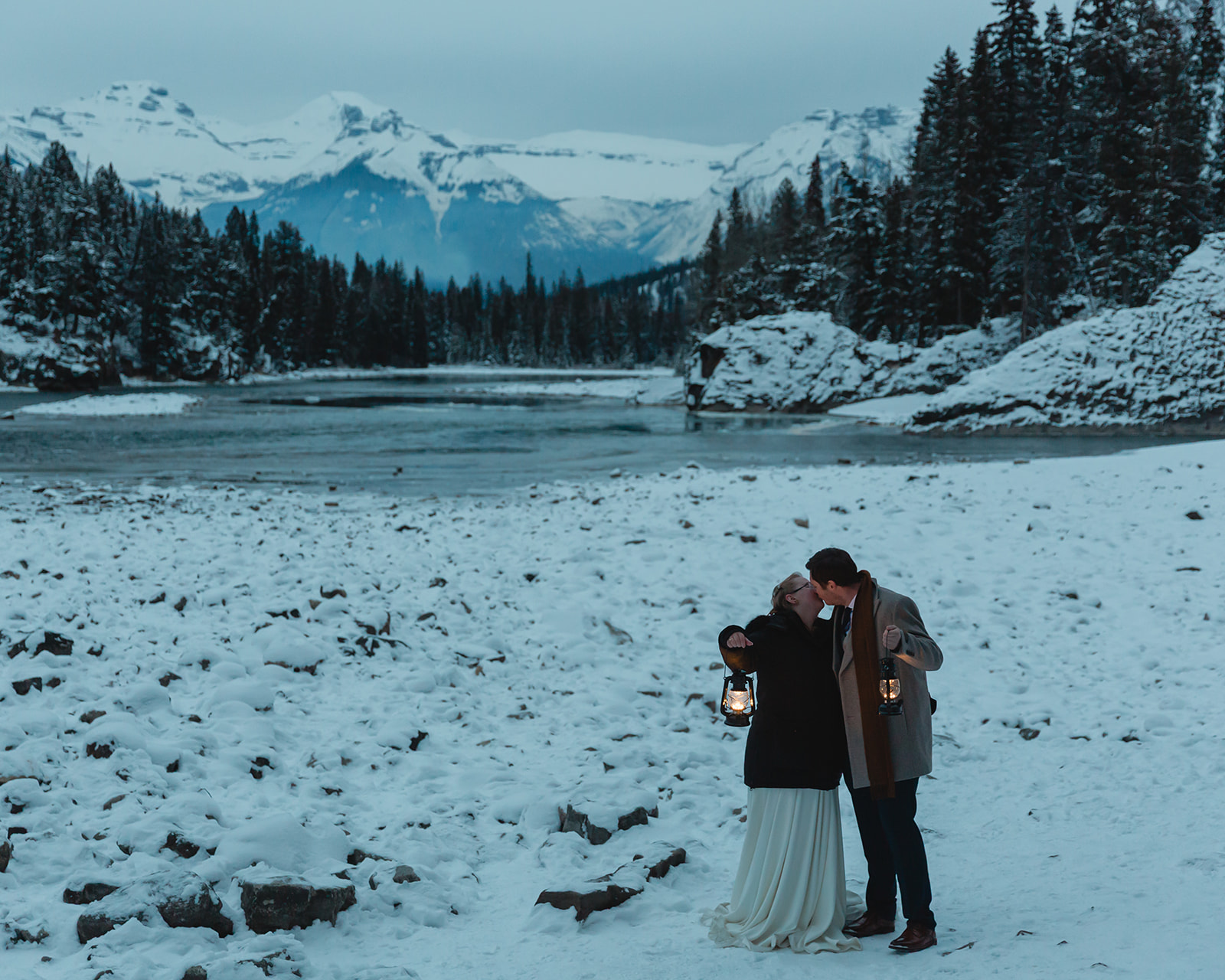 Gabriella and Bronson kiss while holding glowing lanterns at Bow Falls during their snowy Banff winter elopement at blue hour.