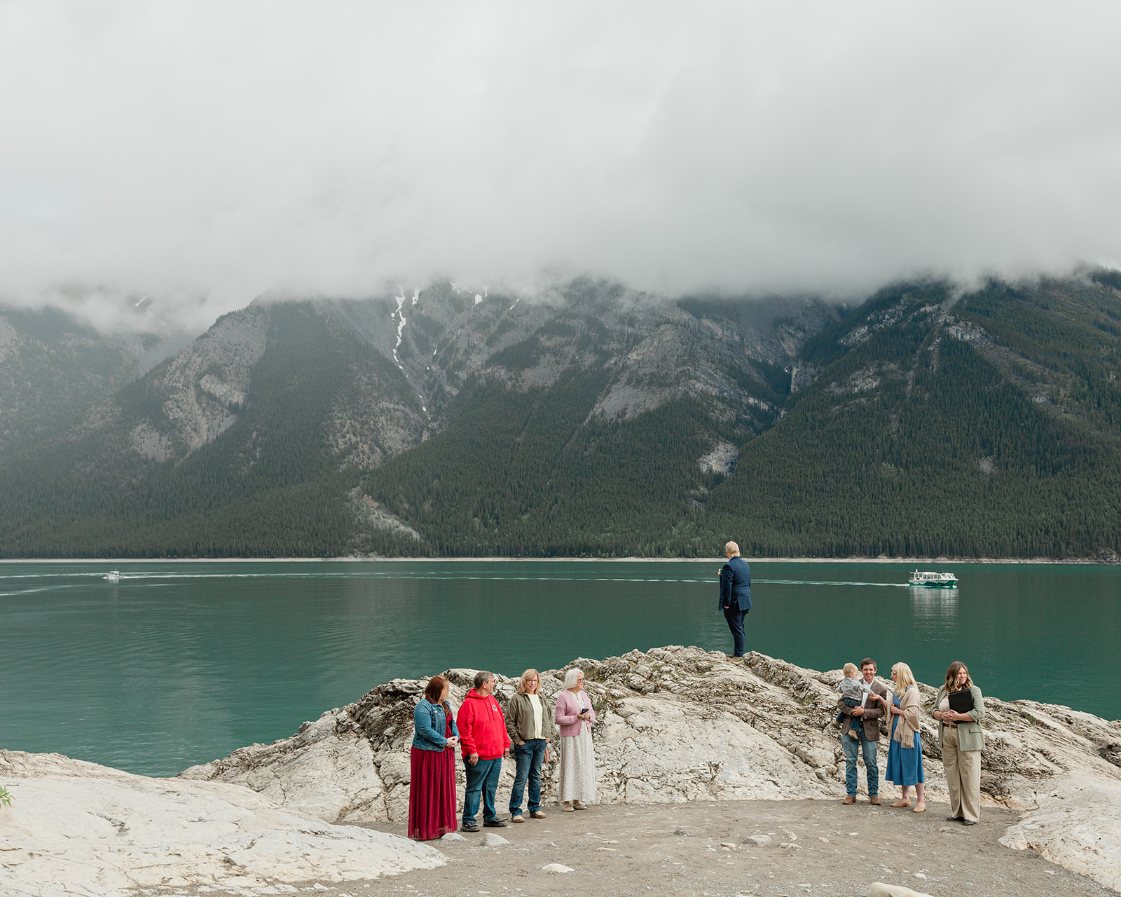 Groom waiting with family on the rocky Lake Minnewanka shoreline beneath misty mountains before his elopement ceremony in Banff.