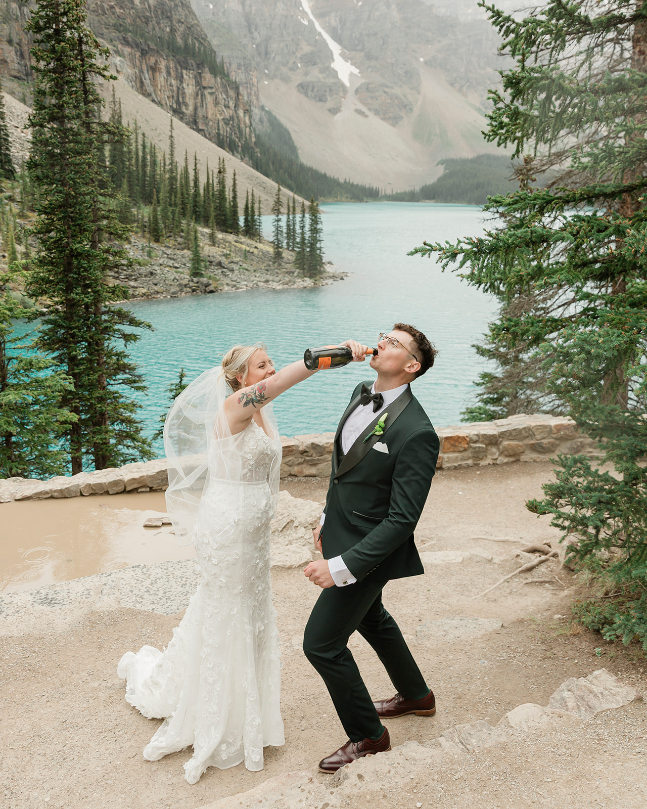 ride playfully pouring champagne into the groom’s mouth during their Moraine Lake elopement, surrounded by turquoise water and the Canadian Rockies.