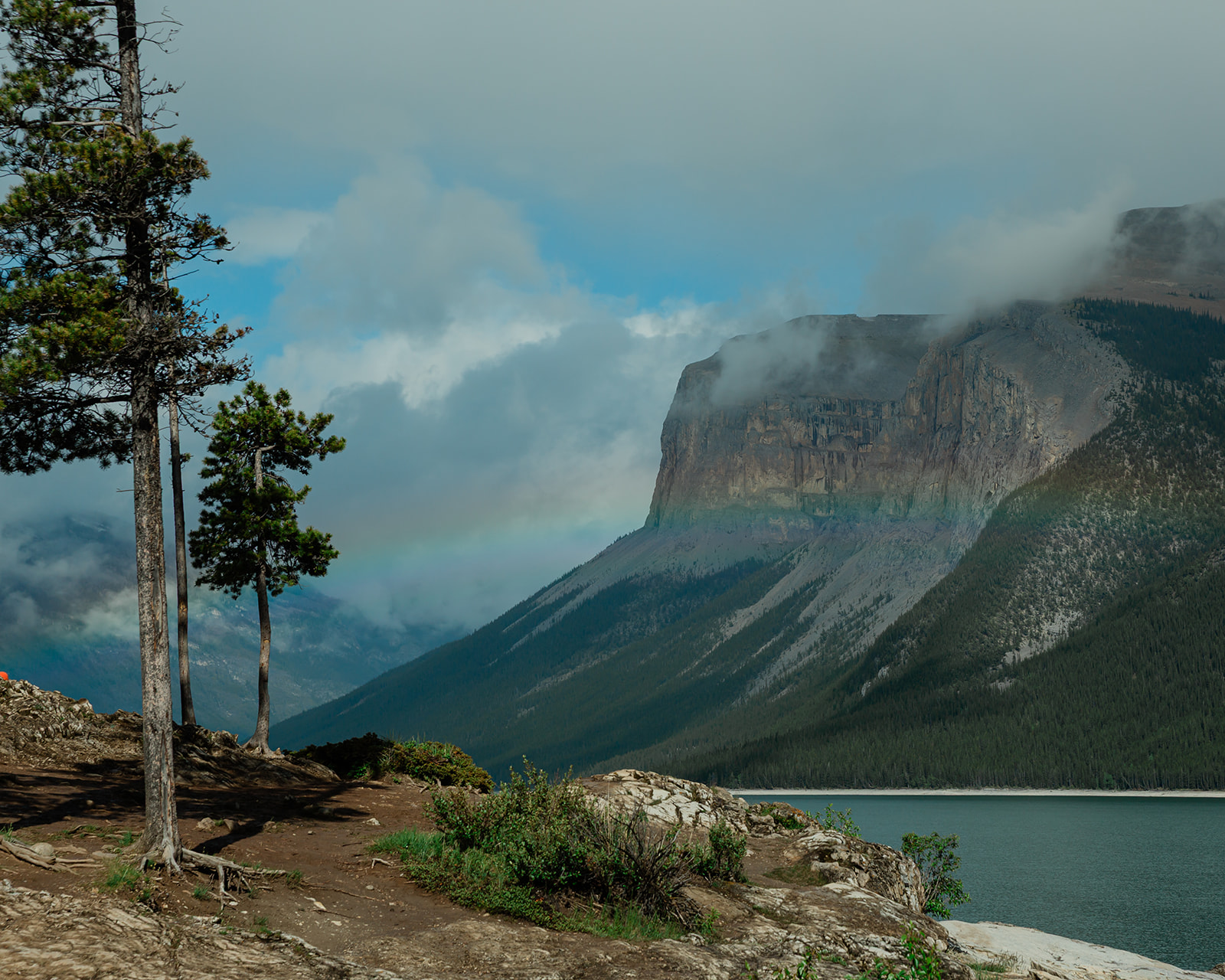 Dramatic mountain landscape at Lake Minnewanka in Banff National Park, with turquoise water, rocky shoreline, pine trees, and low clouds drifting across the Canadian Rockies.