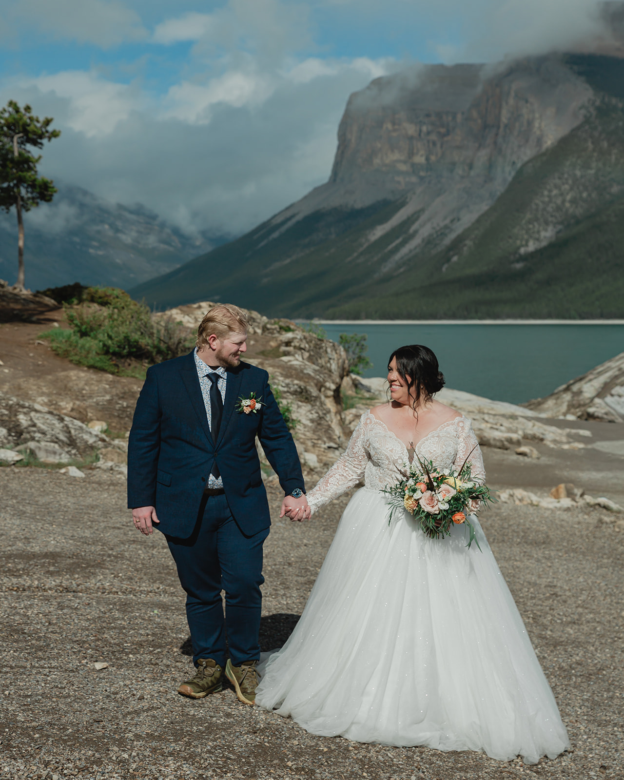 Rachel and Russell walking hand in hand after their Lake Minnewanka elopement, with the turquoise lake and dramatic Canadian Rockies behind them.