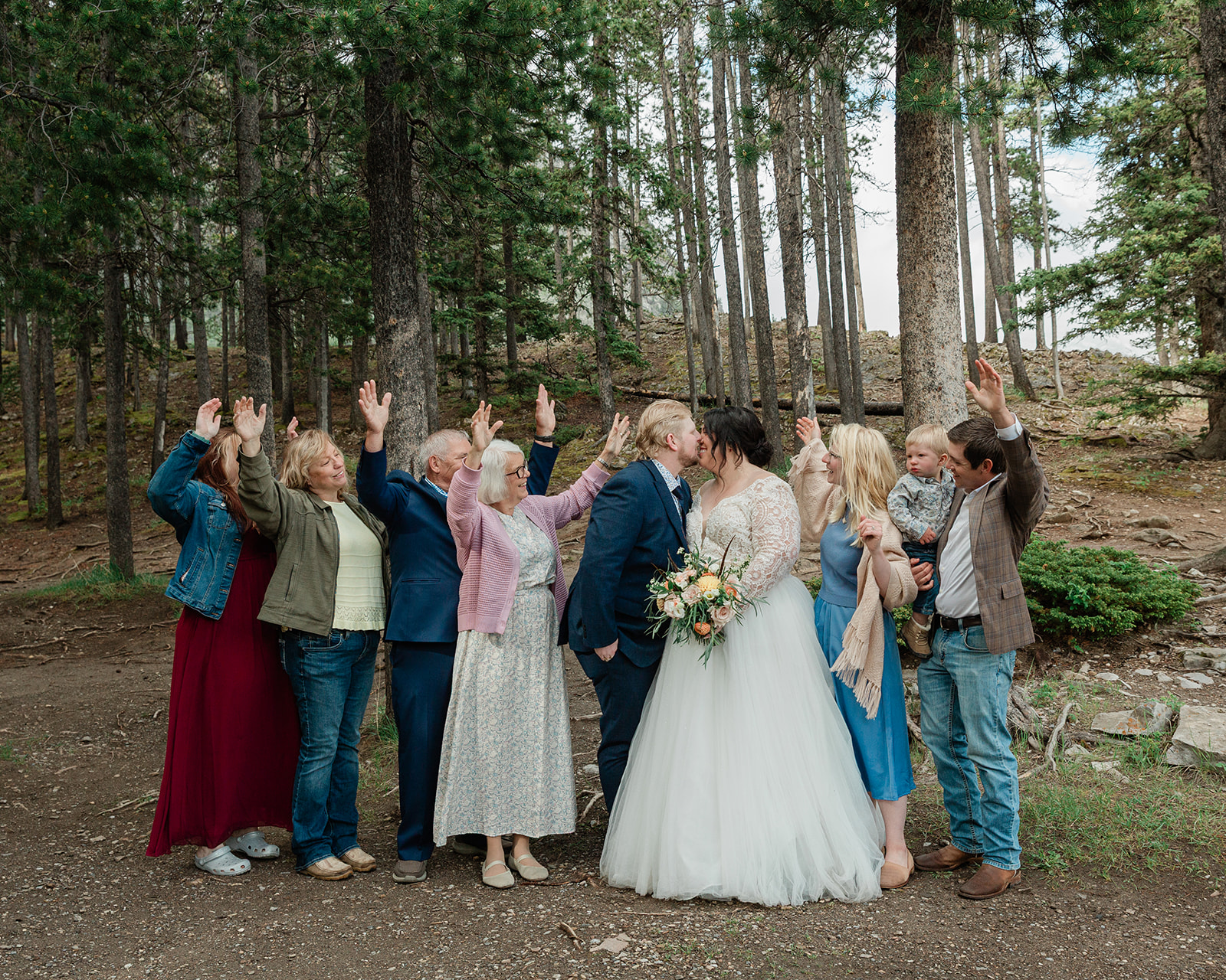 Bride and groom kiss while surrounded by family and loved ones celebrating in a forest near Lake Minnewanka during their intimate Banff elopement.