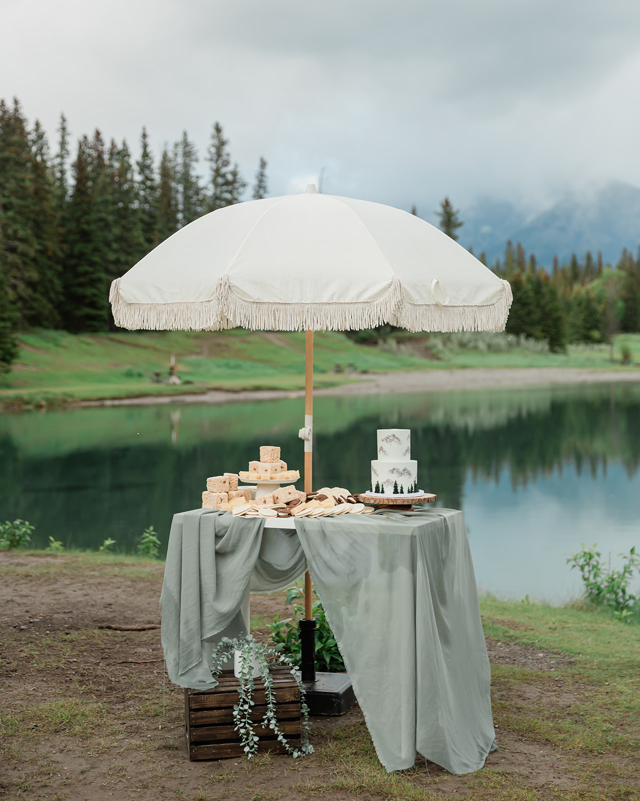 Elegant elopement dessert and snack table beneath a fringed white umbrella beside a mountain lake in Banff National Park.