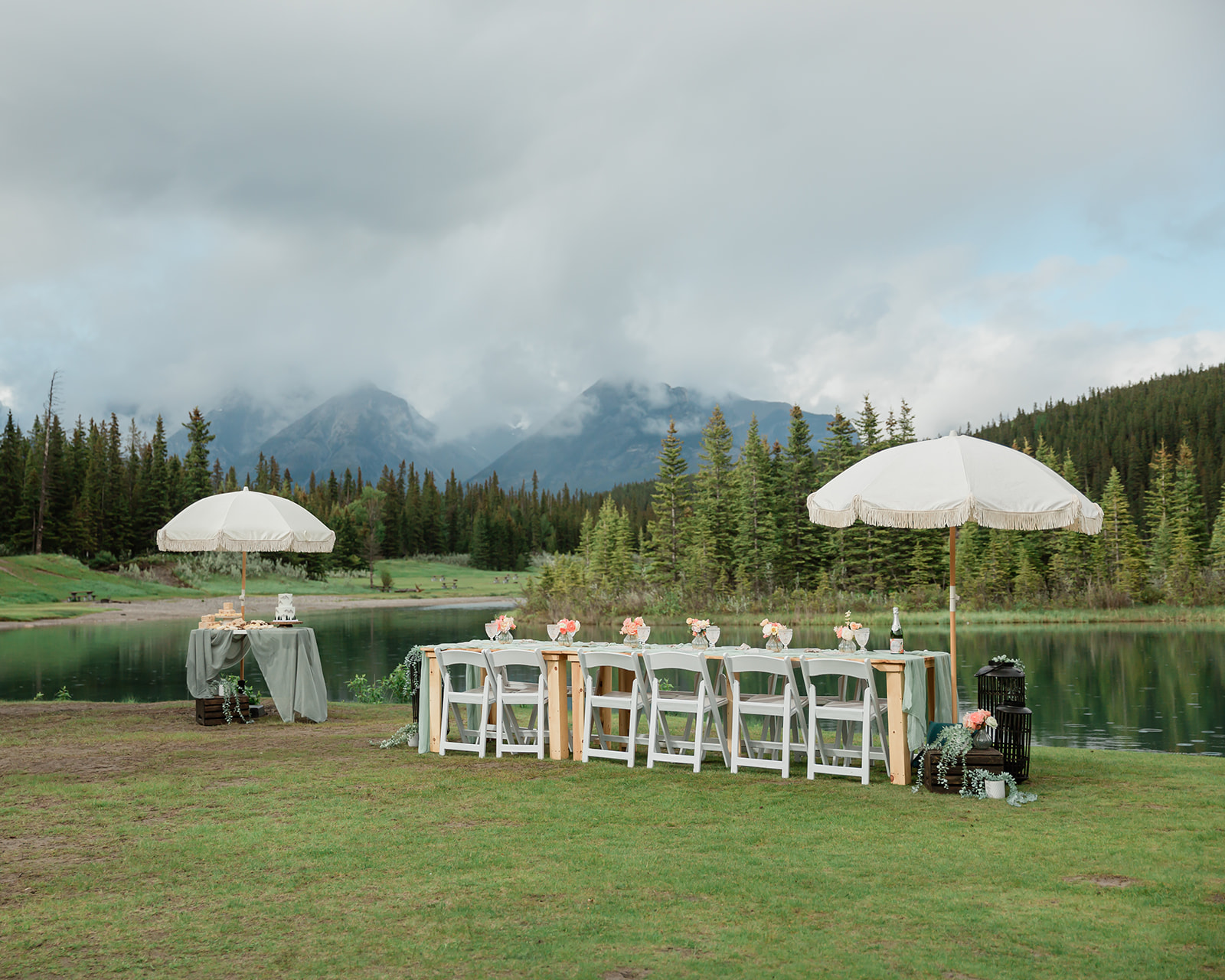 Intimate Banff elopement reception setup with a long outdoor dining table, white chairs, floral details, and fringed umbrellas beside a mountain lake in the Canadian Rockies.