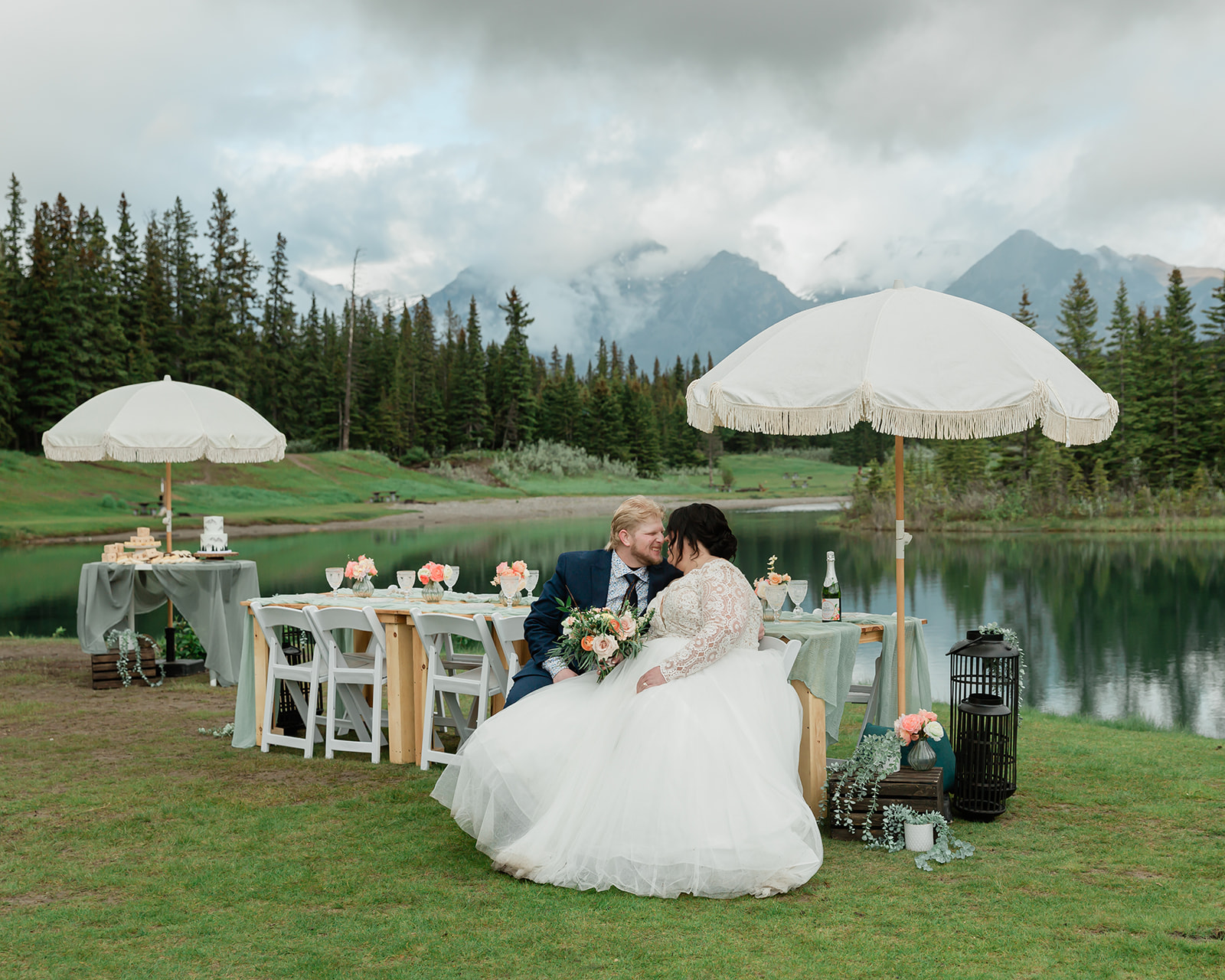 Newlywed couple seated together beside an intimate lakeside reception table with white umbrellas, floral details, and misty Canadian Rocky Mountains in the background during their Banff elopement.