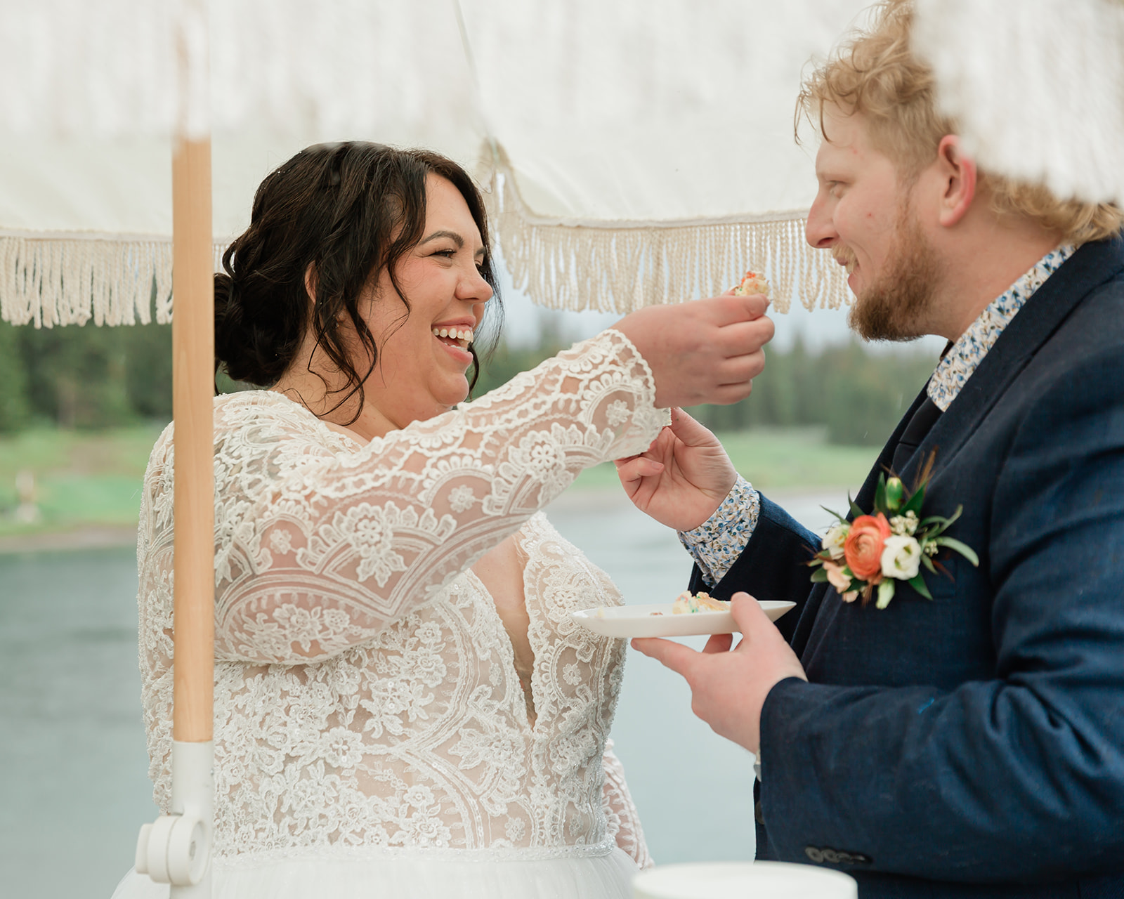 Bride laughing as she feeds cake to her partner beneath a fringed umbrella during their intimate Banff elopement reception, with a mountain lake in the background.
