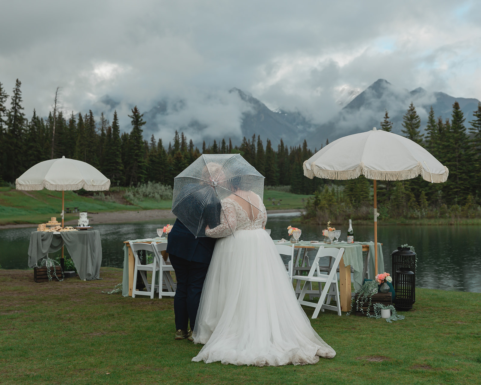 Rachel and Russell share a kiss beneath a clear umbrella during their rainy Banff elopement reception, surrounded by an intimate lakeside table setup and misty mountain views.