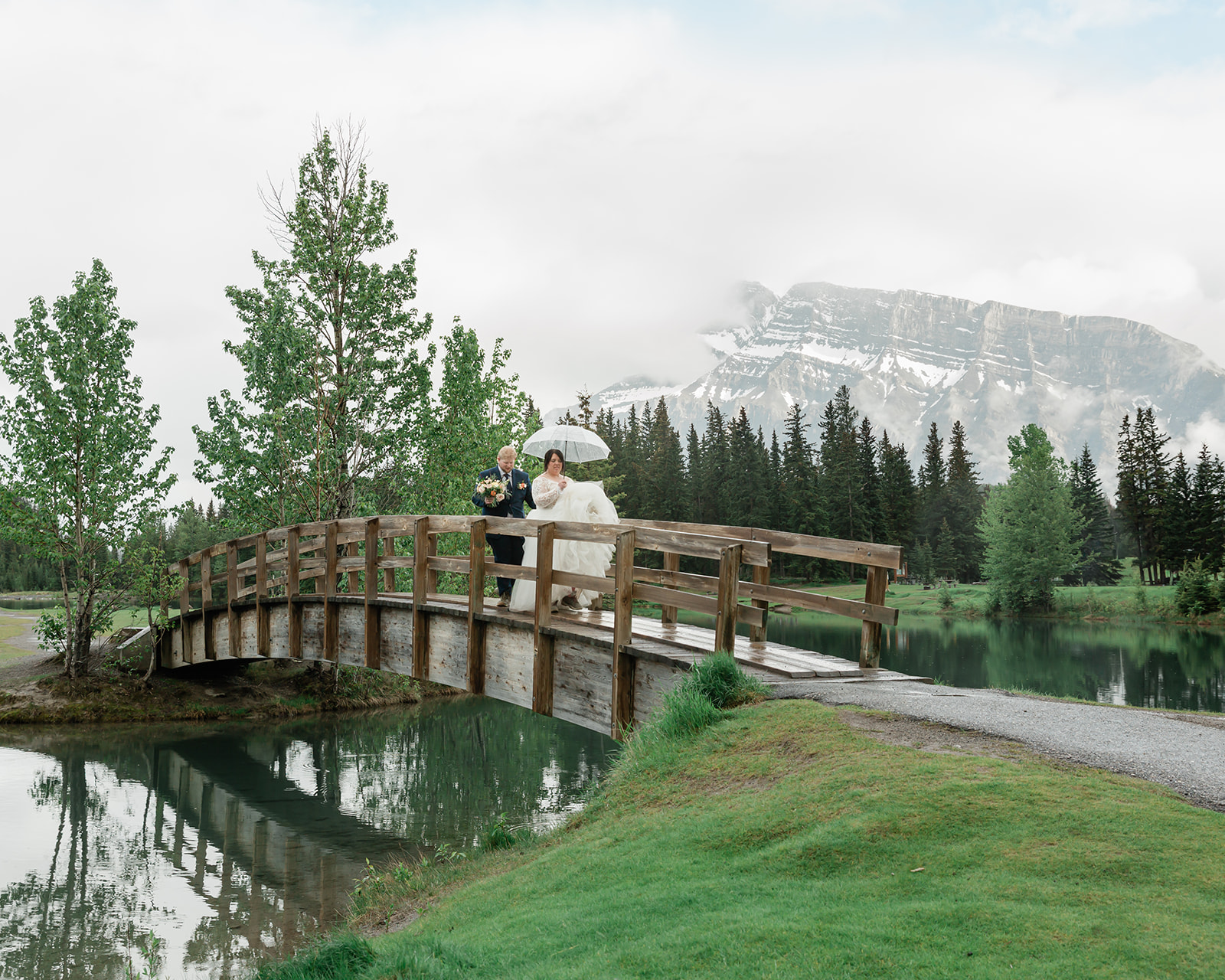 Rachel and Russell walk across a wooden bridge beneath an umbrella during their rainy Banff elopement, with a mountain lake, evergreen forest, and misty Canadian Rockies in the background.