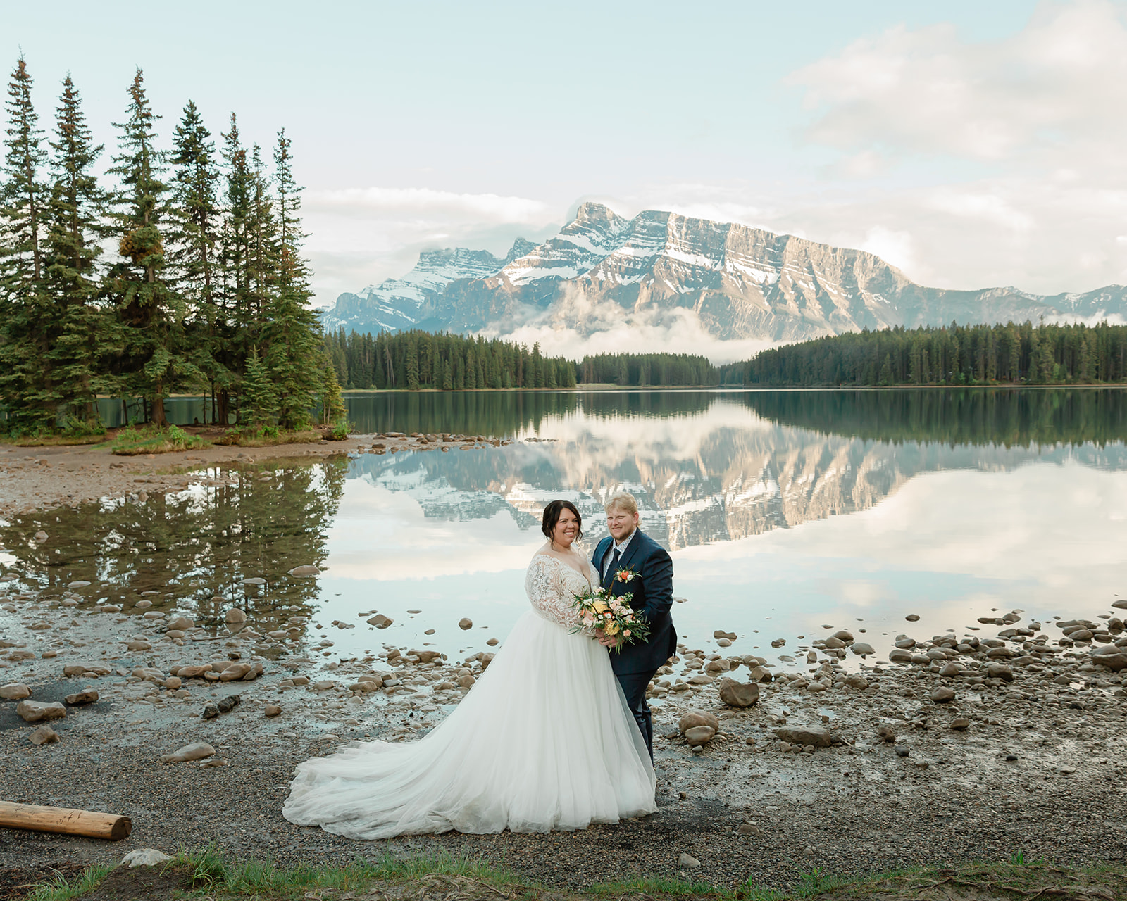 Rachel and Russell stand together on the rocky shore of Two Jack Lake during their Banff elopement, with calm water reflecting the Canadian Rockies and pine forest behind them.