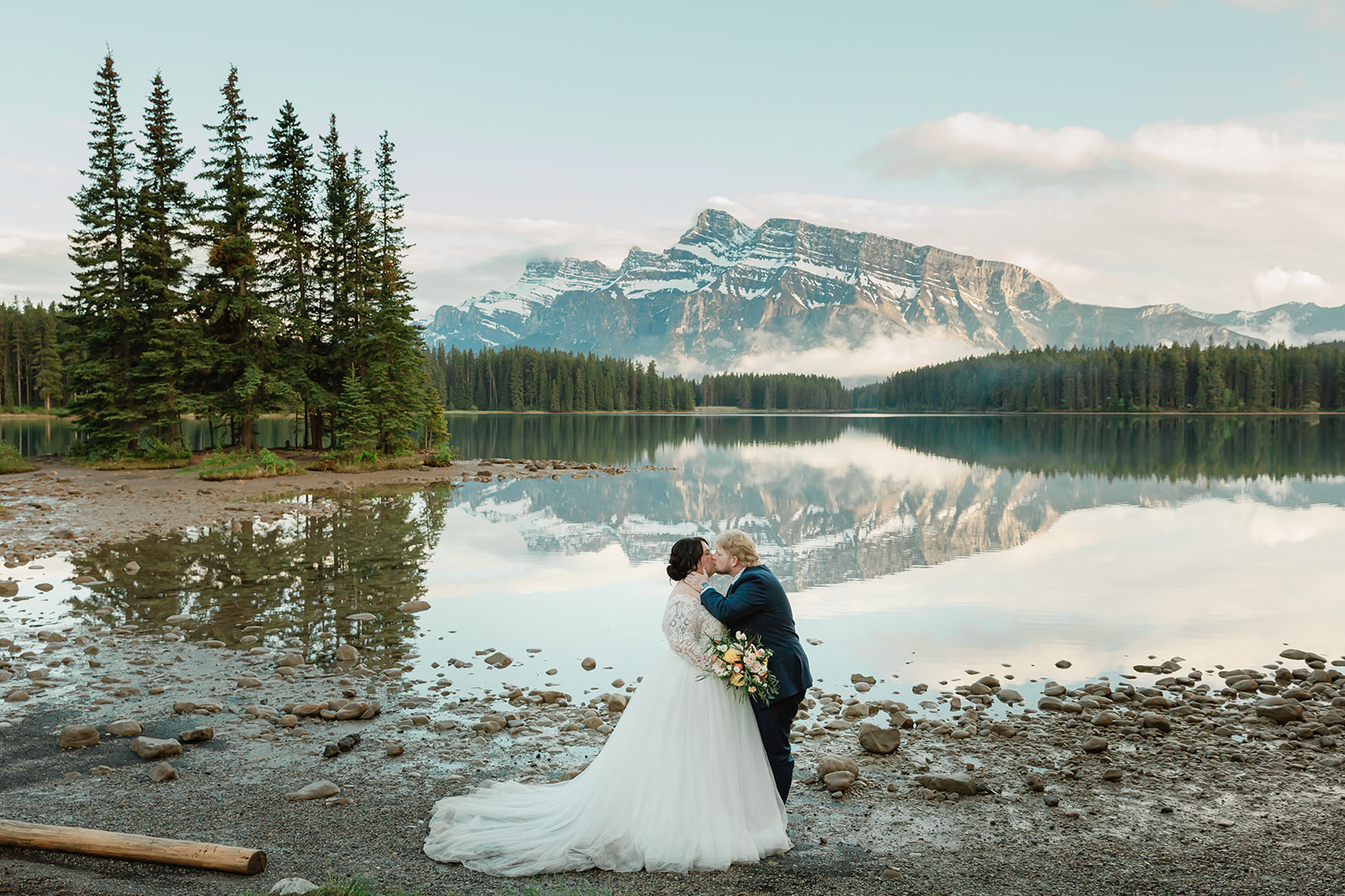 Rachel and Russell kiss along the rocky shore of Two Jack Lake during their Banff elopement, surrounded by calm reflections, evergreen trees, and the Canadian Rockies.