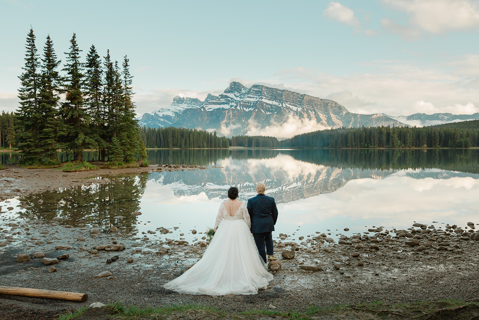Rachel and Russell stand together on the shore of Two Jack Lake during their Banff elopement, looking out at the Canadian Rockies reflected in the calm water.