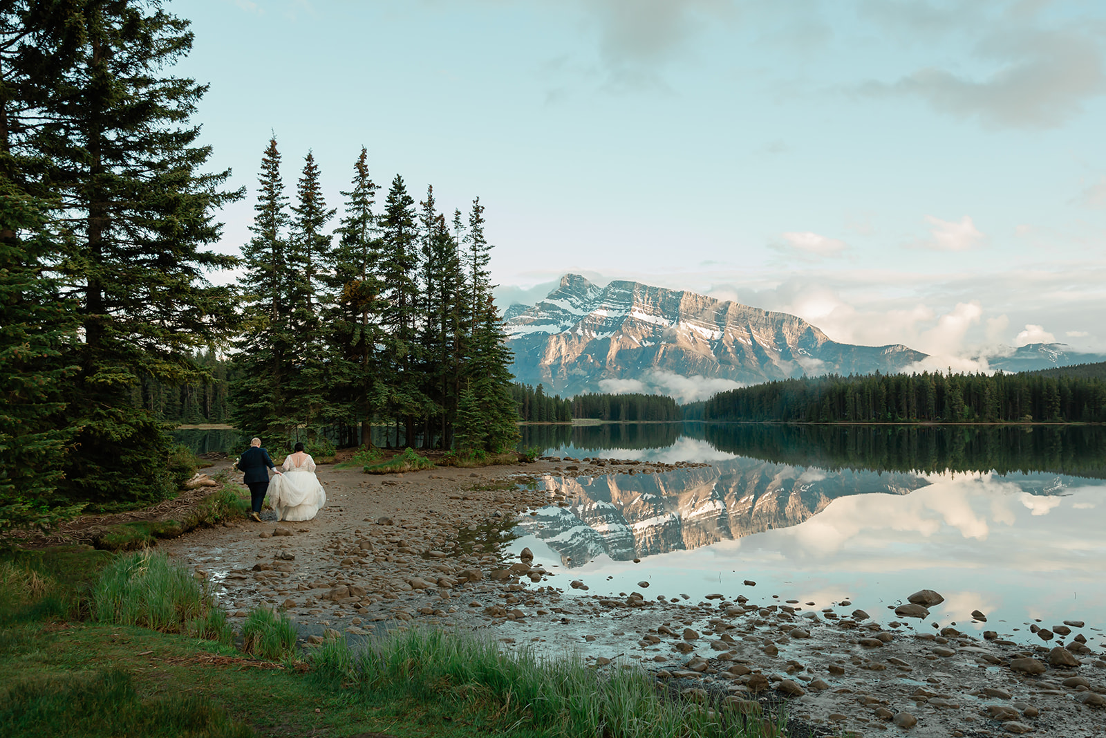 Rachel and Russell walk along the shore of Two Jack Lake during their Banff elopement, surrounded by evergreen trees with the Canadian Rockies reflected in the calm water.