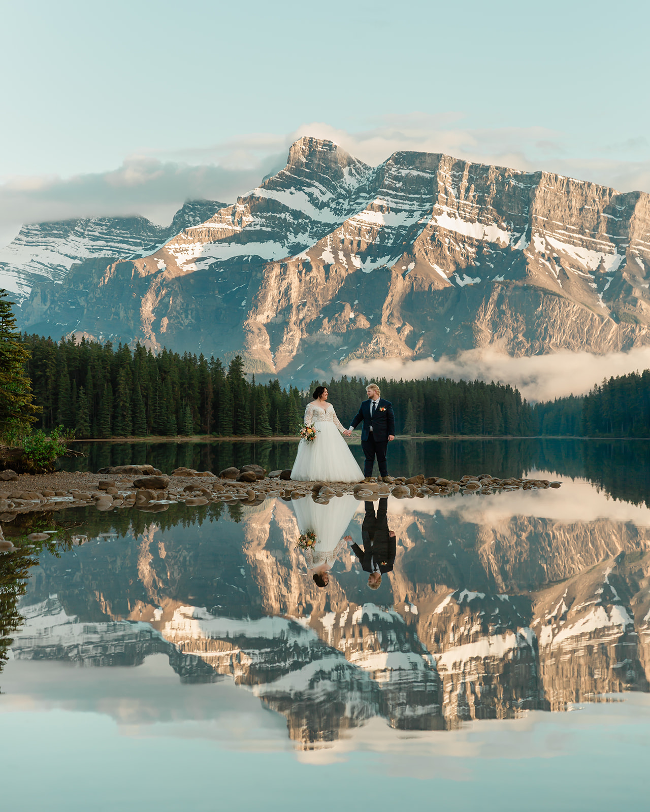 Rachel and Russell stand hand in hand along the shore of Two Jack Lake during their Banff elopement, framed by the Canadian Rockies and their reflection in the calm water.