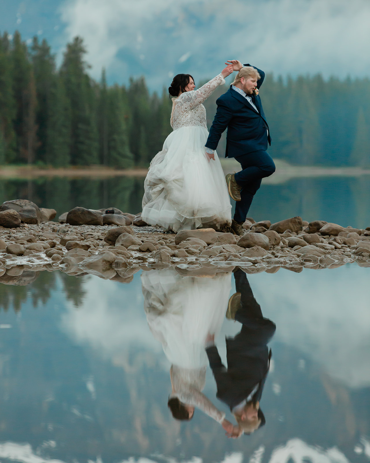 Rachel and Russell playfully dance along the rocky shoreline of Two Jack Lake during their Banff elopement, with their reflection visible in the calm mountain water.