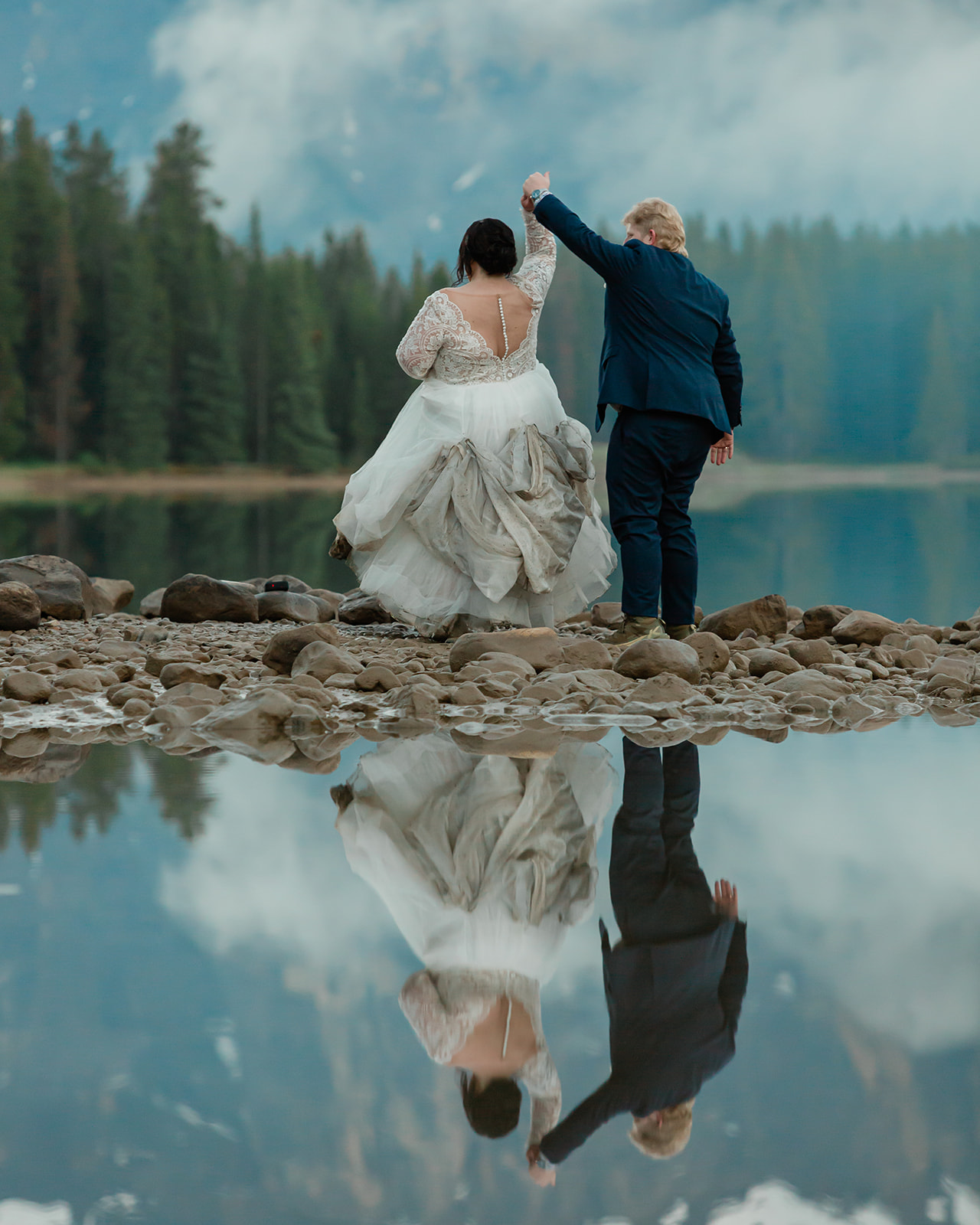 Rachel and Russell twirl together on the rocky shoreline of Two Jack Lake during their Banff elopement, reflected in the calm water with the Canadian Rockies in the background.