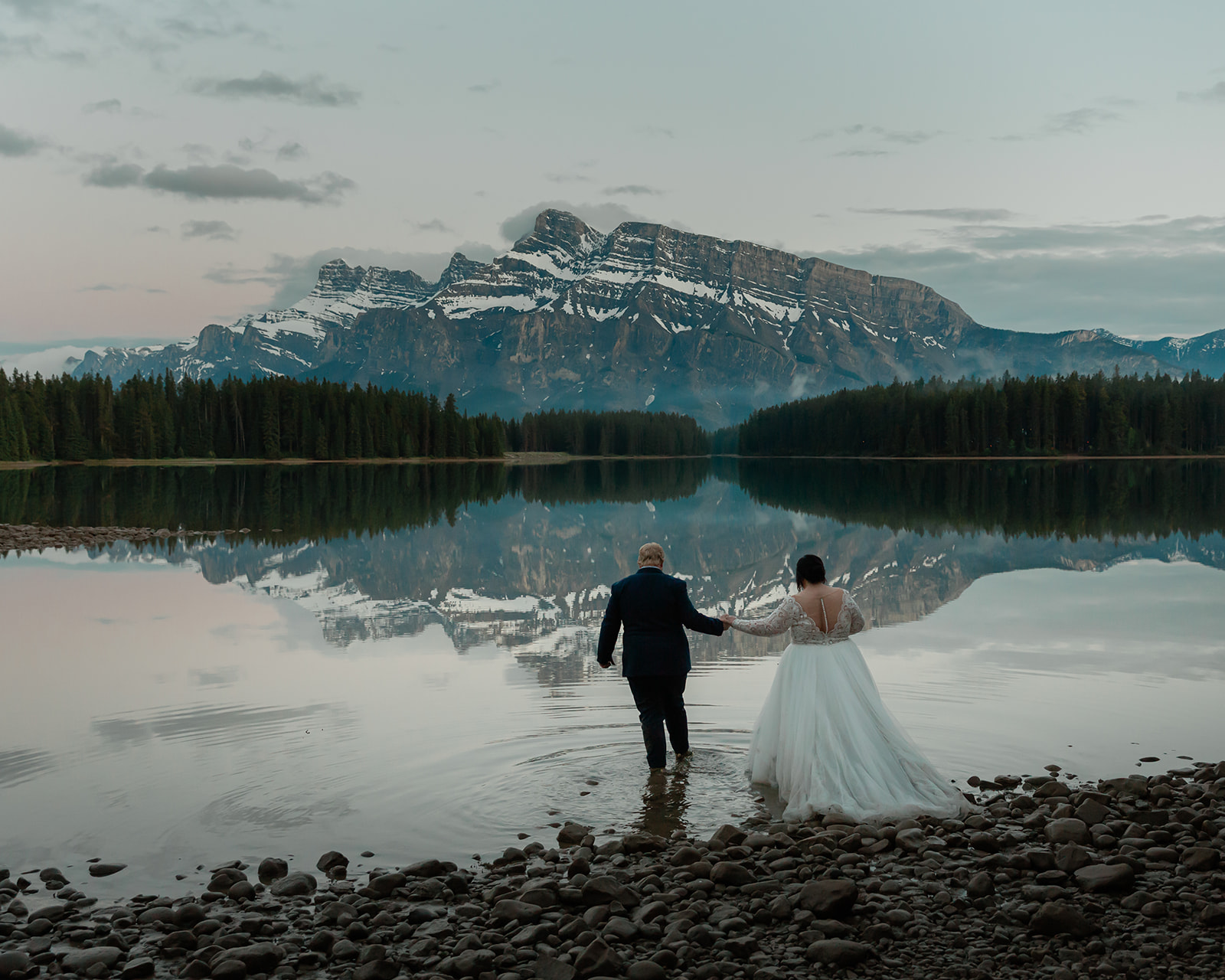 Rachel and Russell hold hands while wading into Two Jack Lake during their Banff elopement, surrounded by calm water reflecting the Canadian Rockies at dusk.